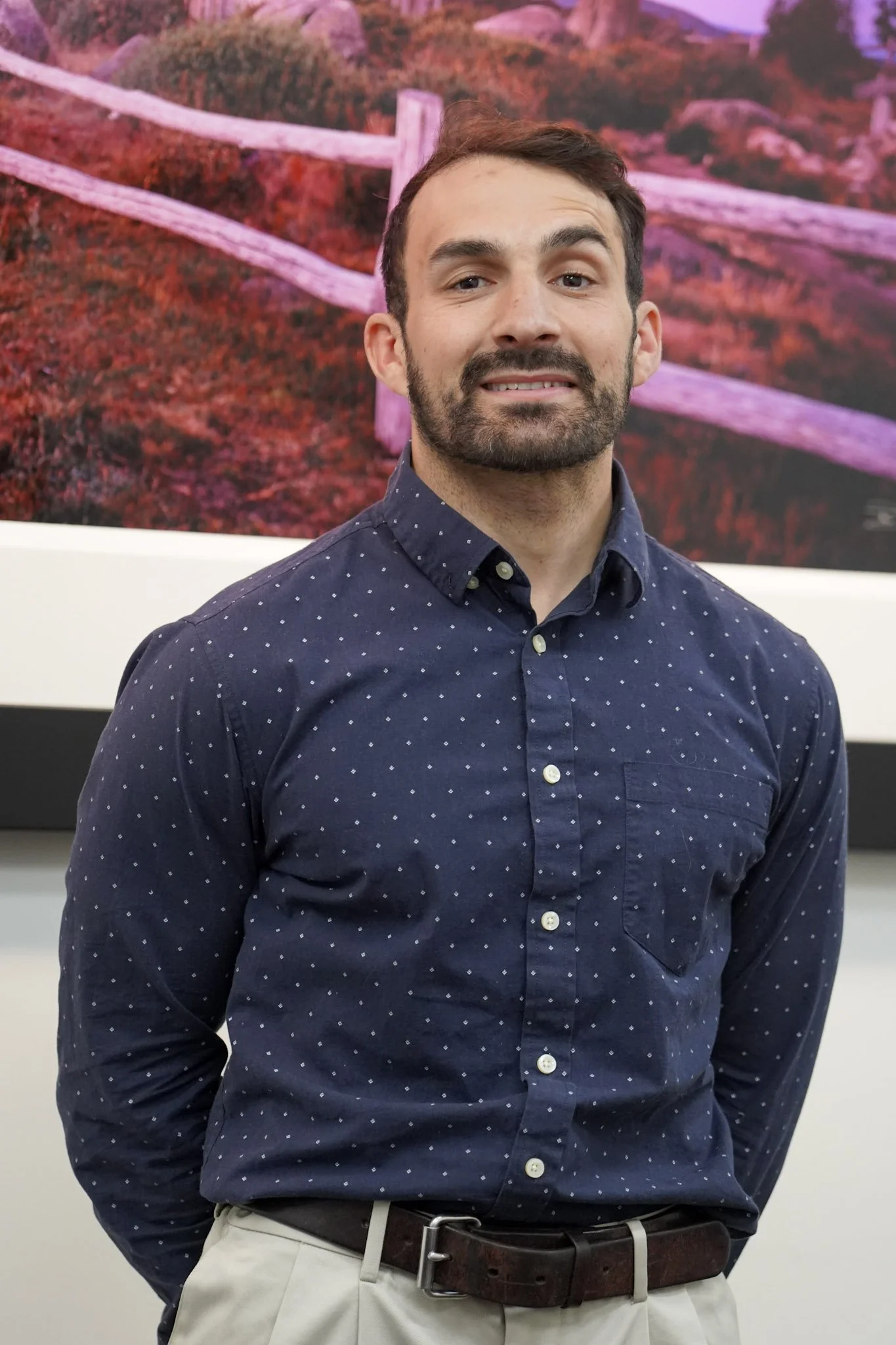 A man with dark hair, a beard, and wearing a navy blue shirt with white polka dots, standing in front of a colorful landscape photograph with pink, purple, and green hues.
