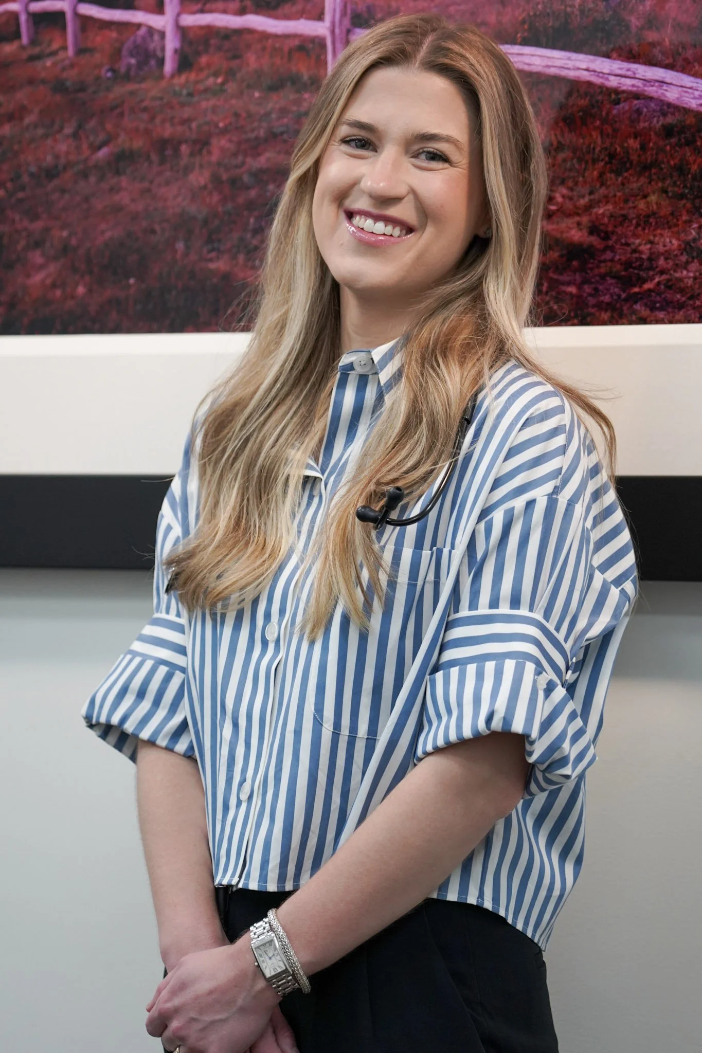 A woman with long, wavy blonde hair is smiling and standing indoors in front of a framed picture of a landscape with pinkish and reddish foliage. She is wearing a blue and white striped shirt, a wristwatch, and has a black headset around her neck.