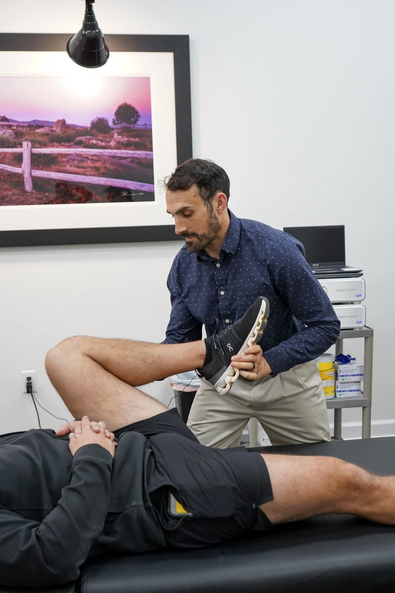 A man lying on an examination table while a healthcare professional holds his lifted leg during a medical or physiotherapy session in a clinical setting.