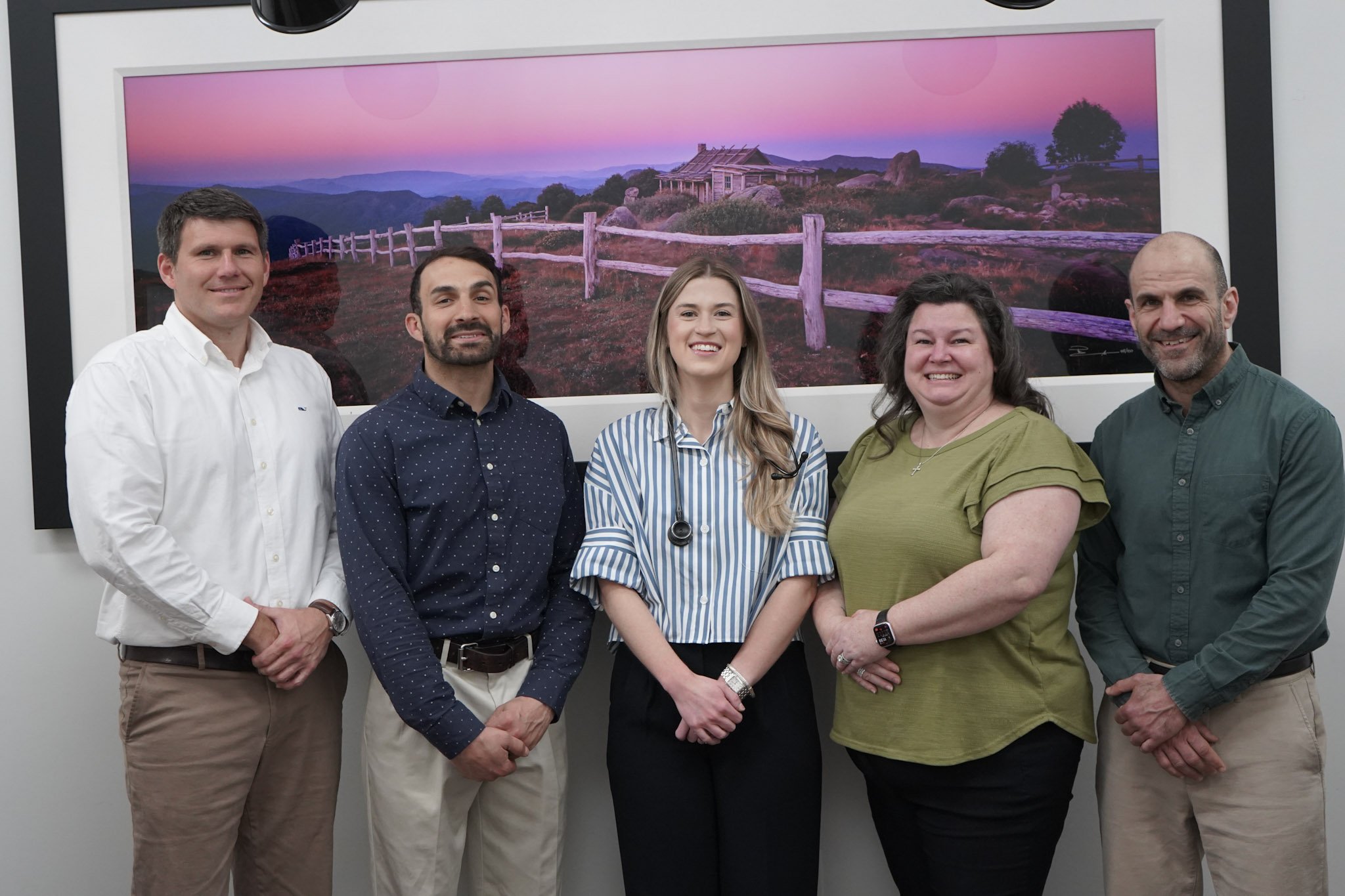 Group of five people standing in front of a large landscape photograph of a sunset over hills, with a house and fence in the background.