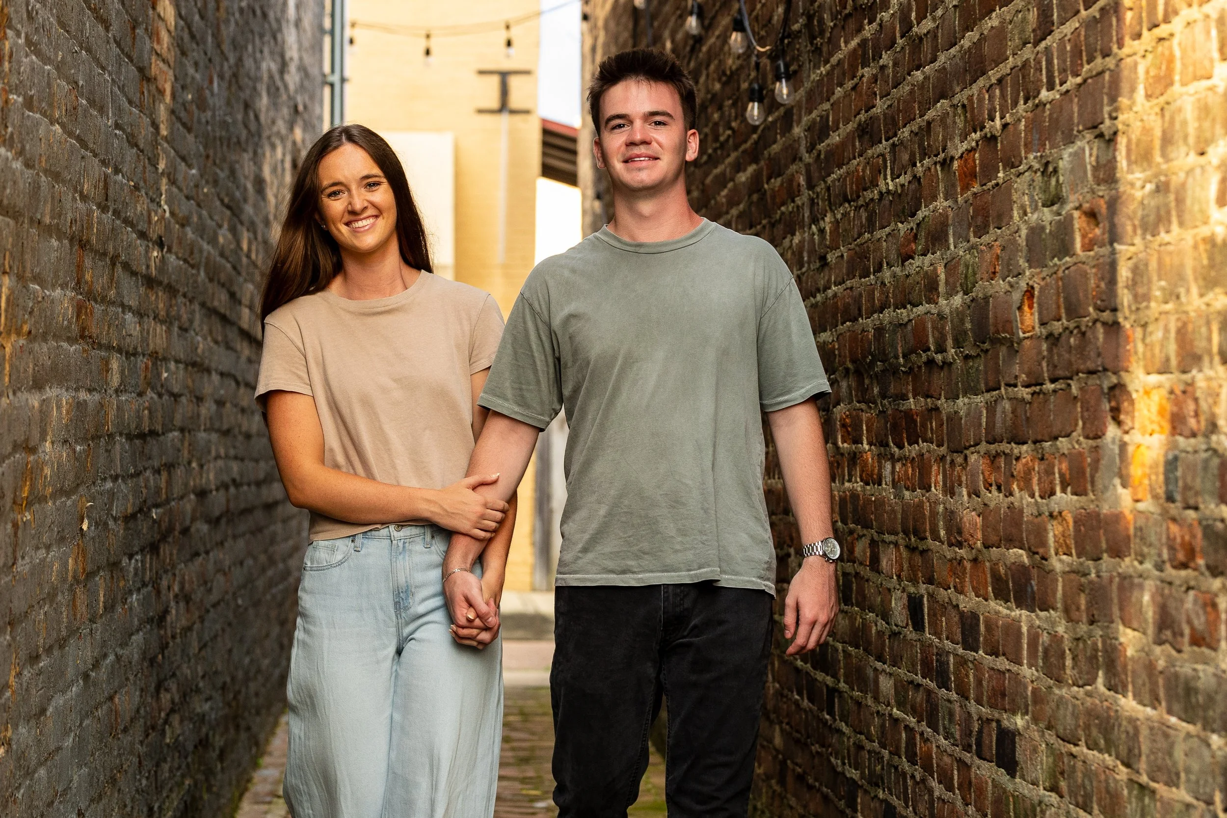 A young couple holding hands and smiling, walking through a narrow alleyway with brick walls on both sides.