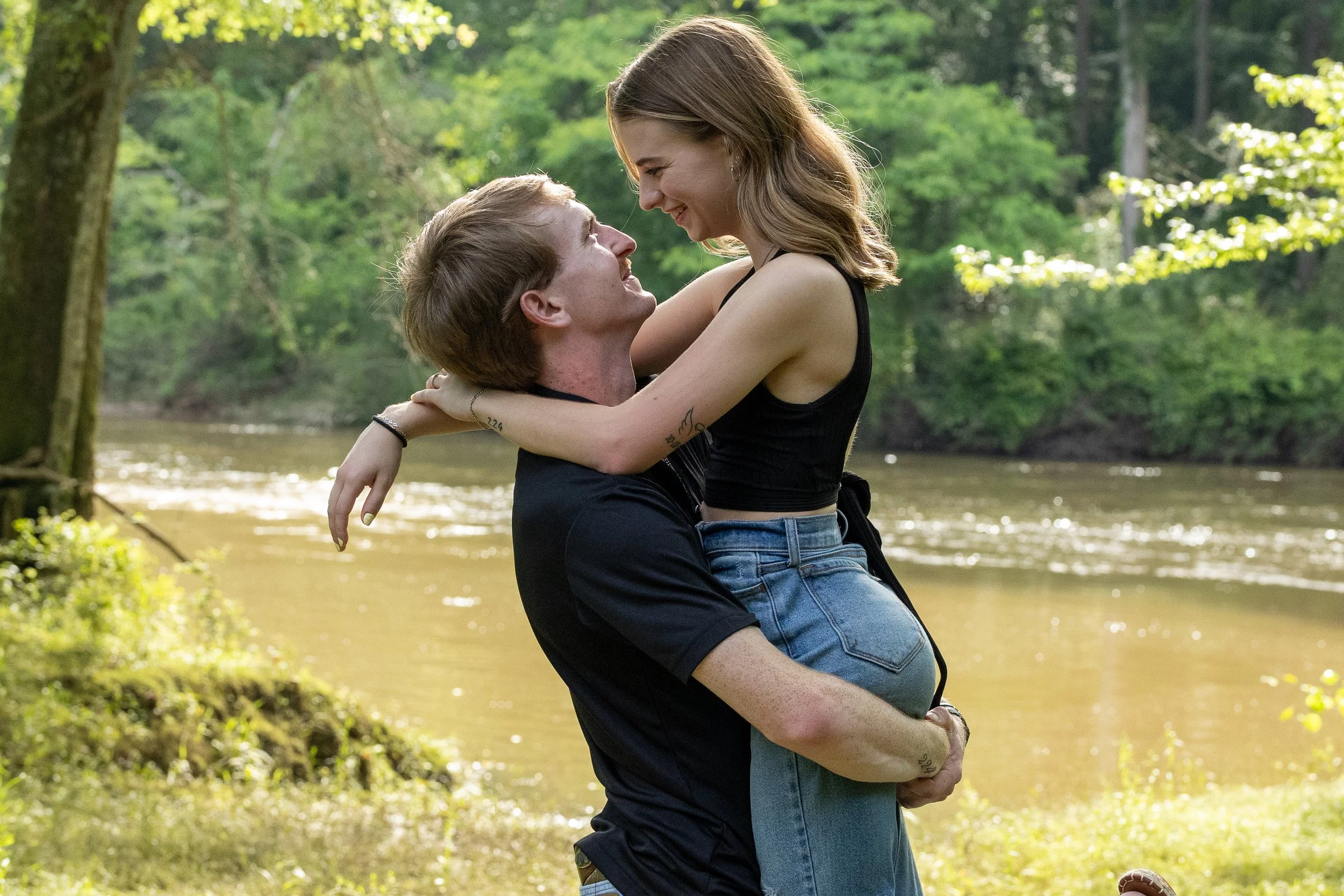A young couple enjoying a moment together outdoors by the river, with the man lifting the woman in his arms as they smile at each other amidst lush greenery.