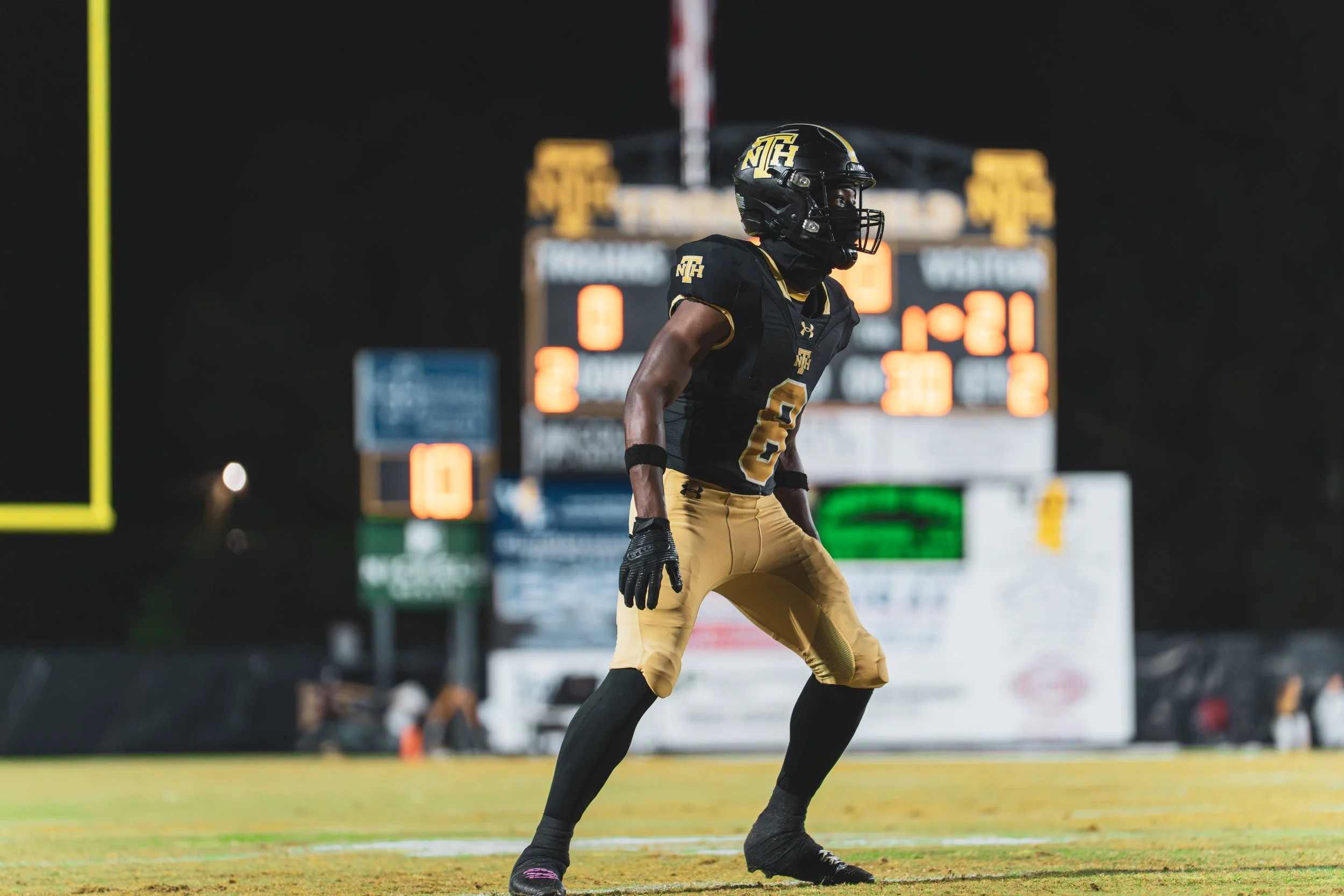Football player on field wearing black and gold uniform, standing with knees bent, number 2, helmet with 'NTH' logo, scoreboard and goalpost in background at night.