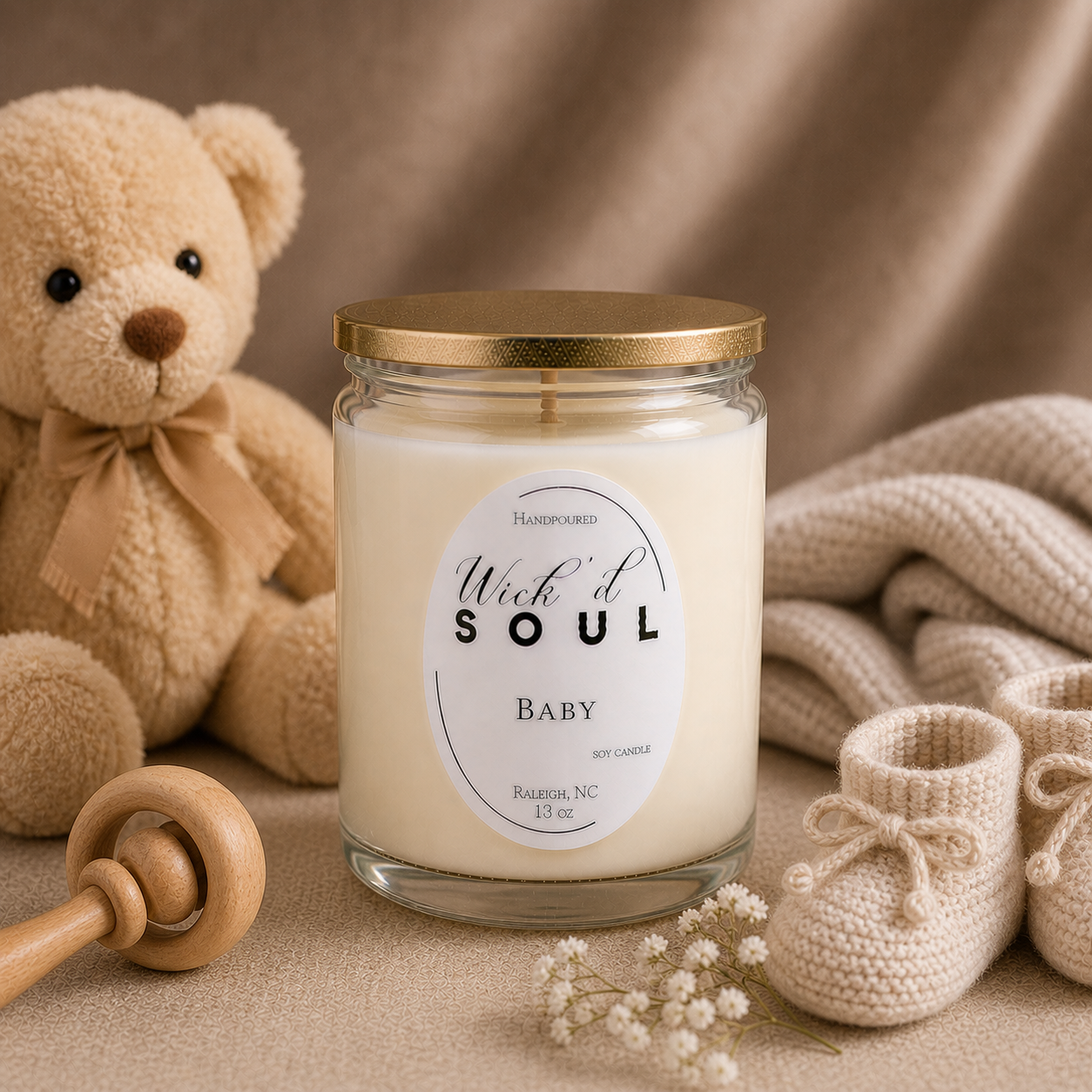 A white soy candle labeled 'Wick'd Soul Baby,' in a glass jar with a gold lid, surrounded by a plush teddy bear with a brown bow, beige knitted baby shoes, a wooden rattle, small white flowers, and soft beige fabric in the background.