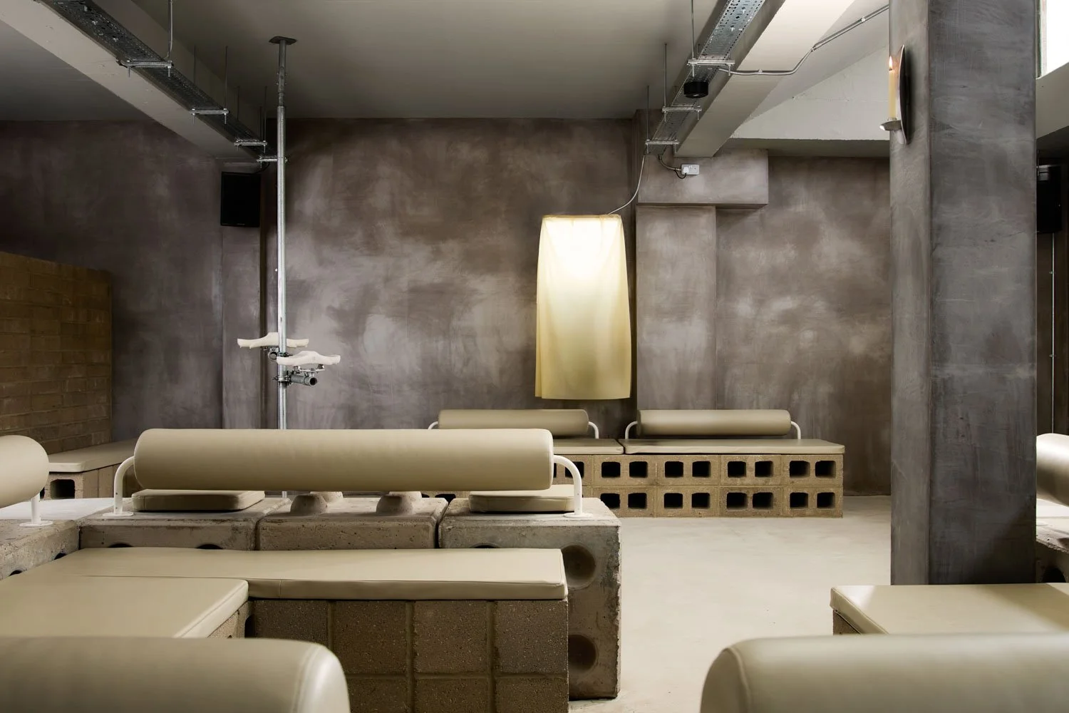 Minimalist interior with concrete walls and cushioned benches, industrial-style lighting, and a tall latex lamp.