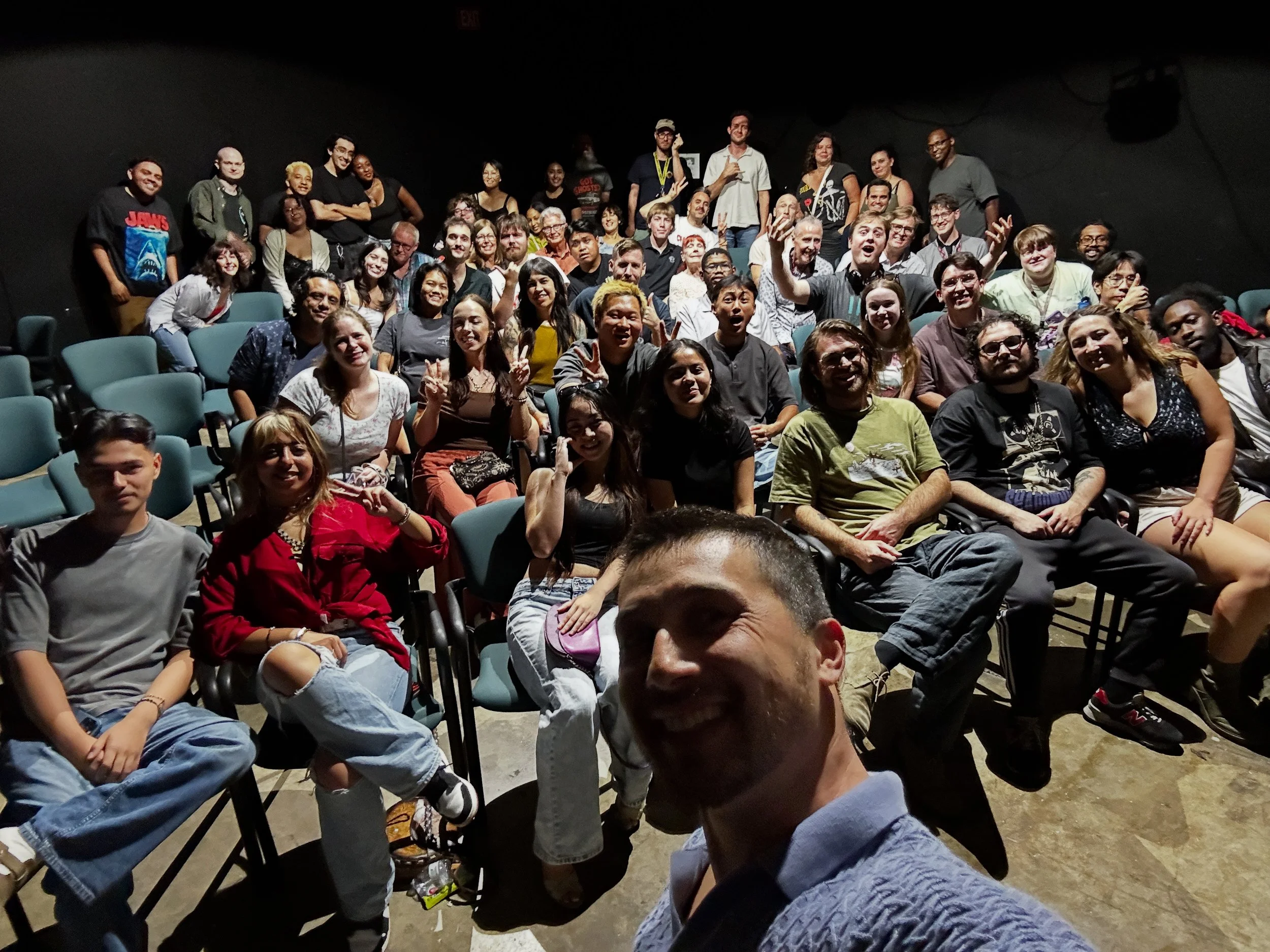 A diverse group of filmmakers and audience members smile and pose for a group photo inside a small theater following a community film screening.