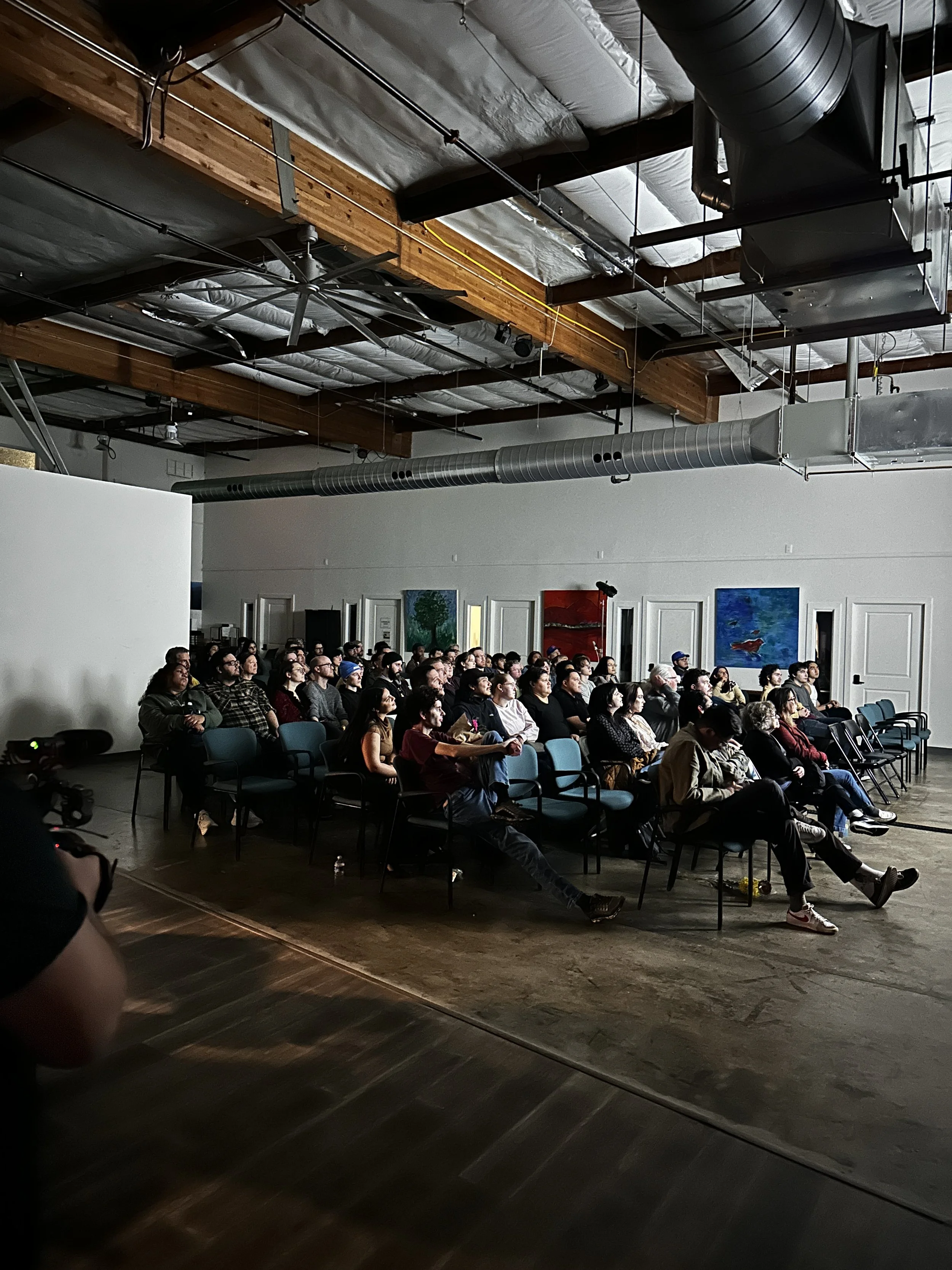 Audience members sit in rows of chairs inside an industrial-style event space, attentively watching a projected film during a screening event.
