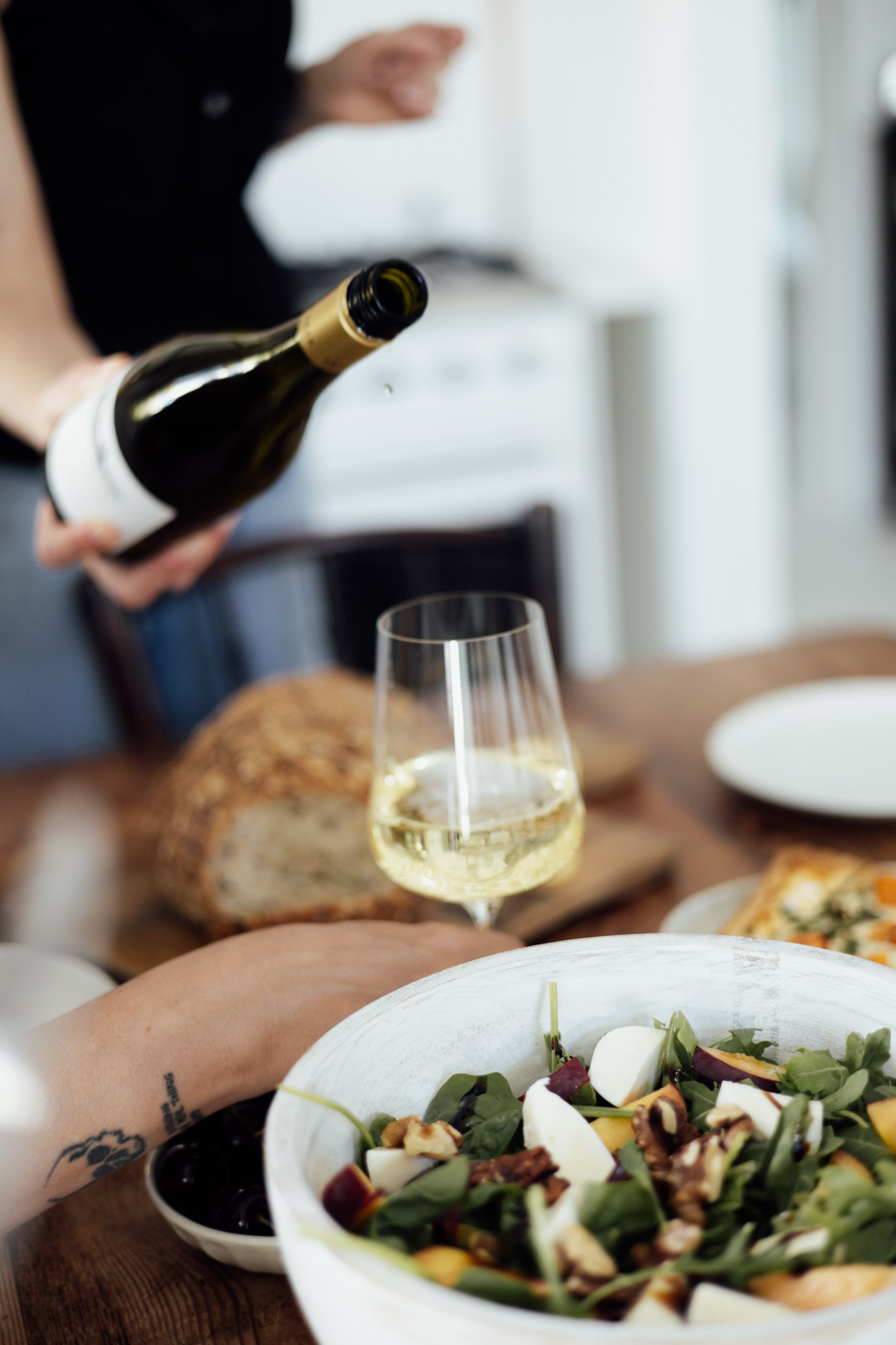 Person pouring white wine into a glass on a dining table with salad, bread, and other dishes.