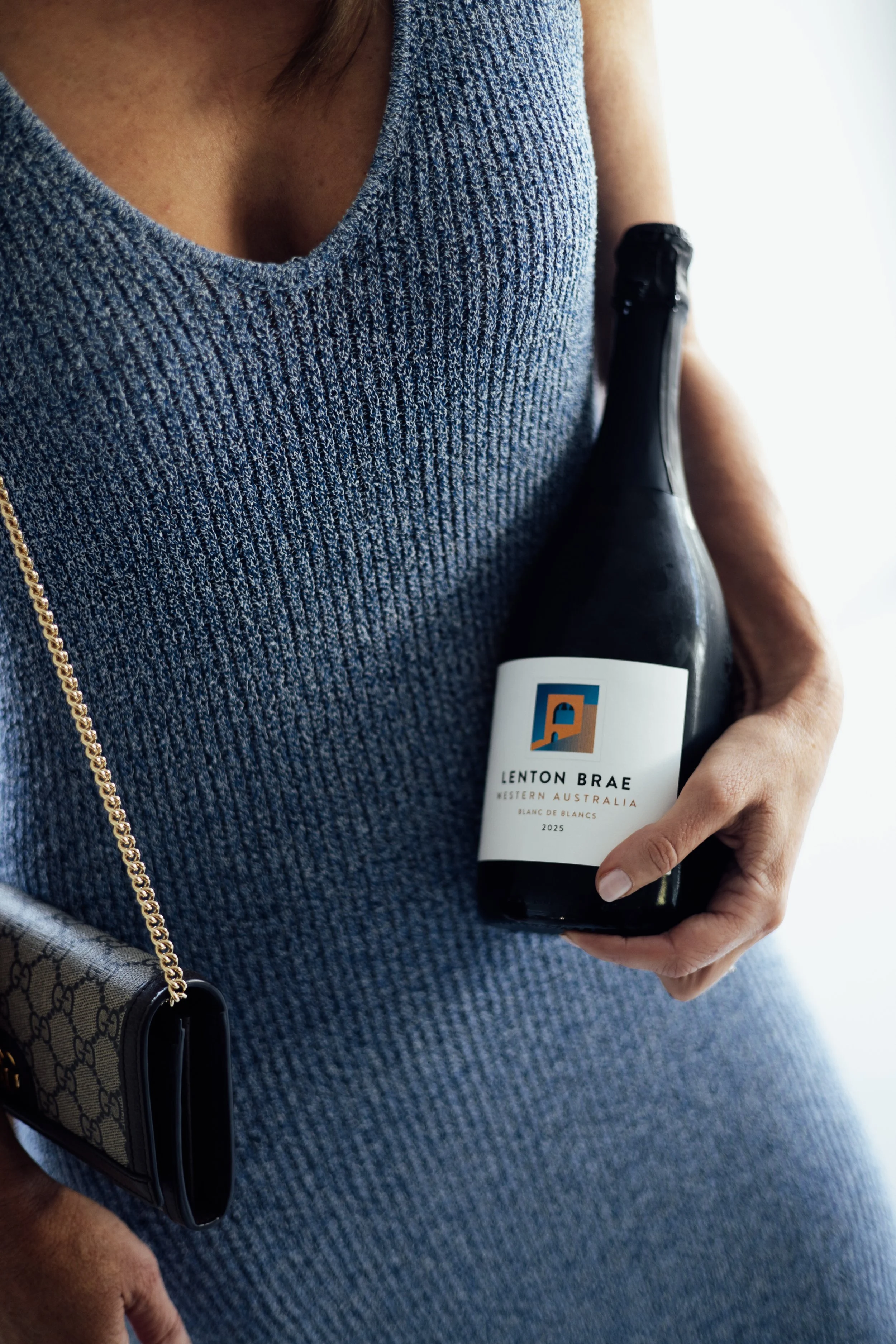 A person wearing a textured blue dress holding a bottle of Lenton Brae wine from Western Australia, vintage 2025, with a small Gucci handbag hanging from their shoulder.