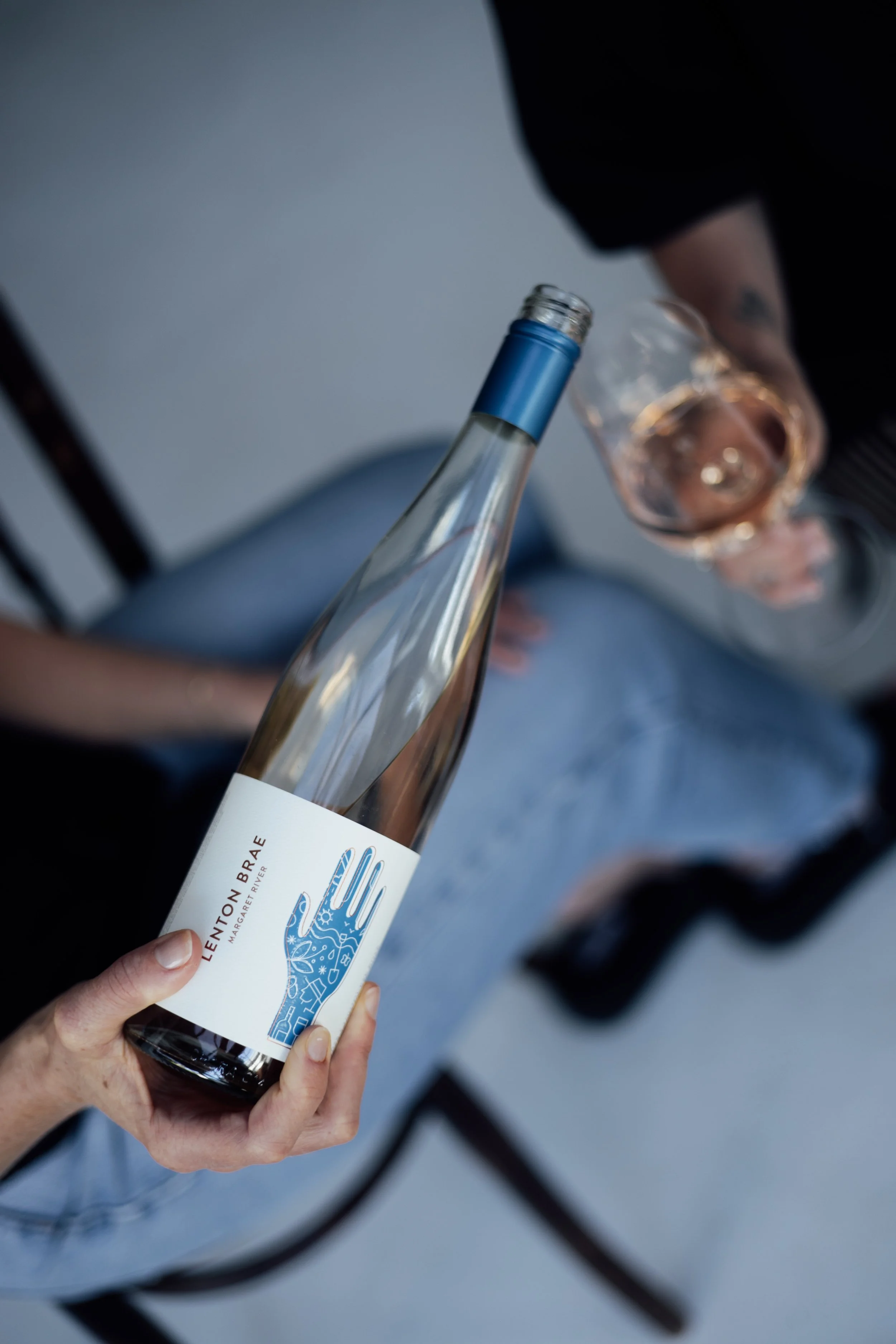 A person holding a bottle of wine labeled 'Lenton Brae' in one hand and a glass of rosé wine in the other, sitting on a chair.