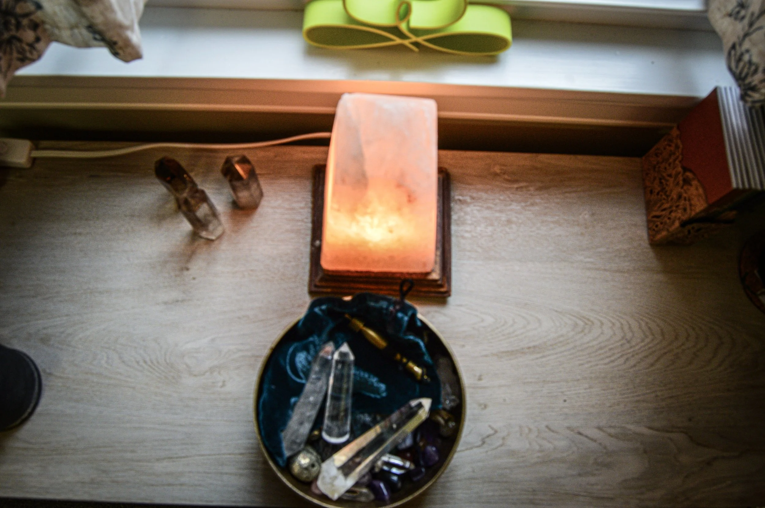 A shelf with a glowing salt lamp, three smaller crystals, a bowl with various crystals and a crystal wand, a green yoga strap, and some books.