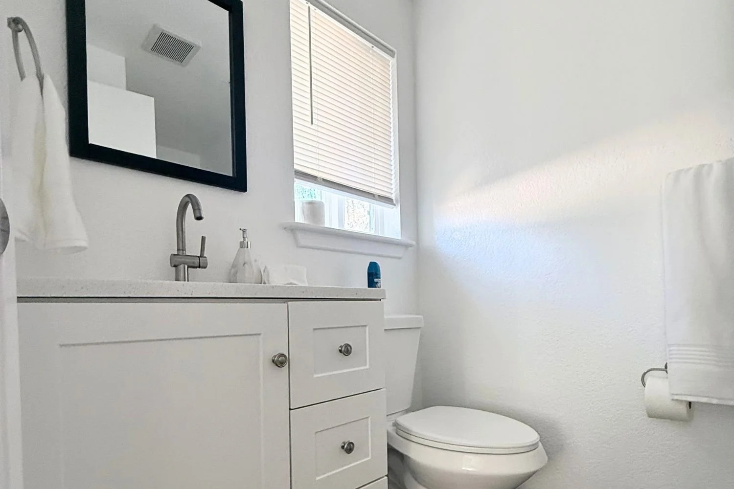 A small white bathroom with a cabinet, mirror, and window with blinds, next to a toilet with a roll of toilet paper on the wall.