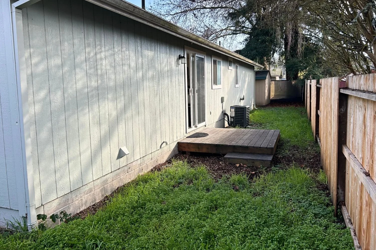 Backyard view of a house with beige siding, small wooden deck, sliding door, and green lawn area, enclosed by a wooden fence, with trees in the background.