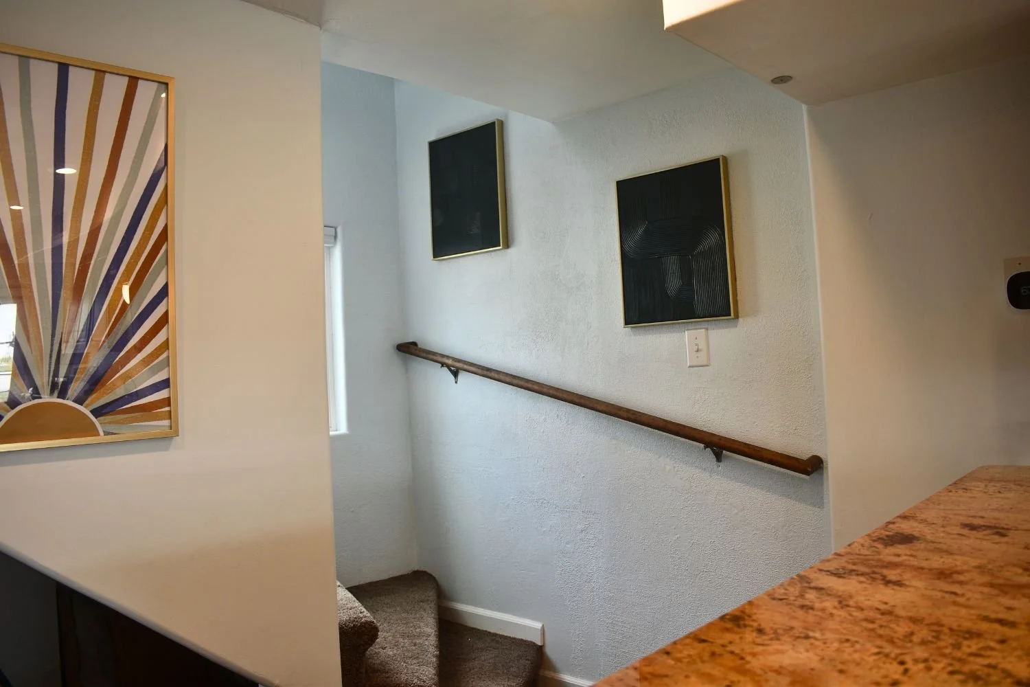 Interior view of a staircase landing with a wooden handrail, two black framed abstract artworks on a white textured wall, a small window, and part of a colorful wall hanging on the left.