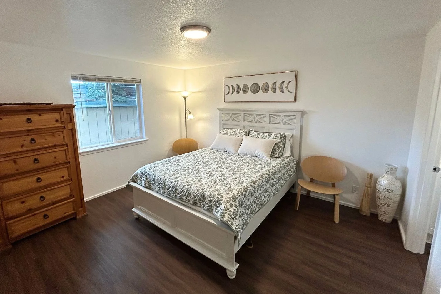 A bedroom with a wooden dresser, a window, a white bed with floral bedding, wall art with moon phases, a lamp, two wooden chairs, and a decorative vase in the corner.