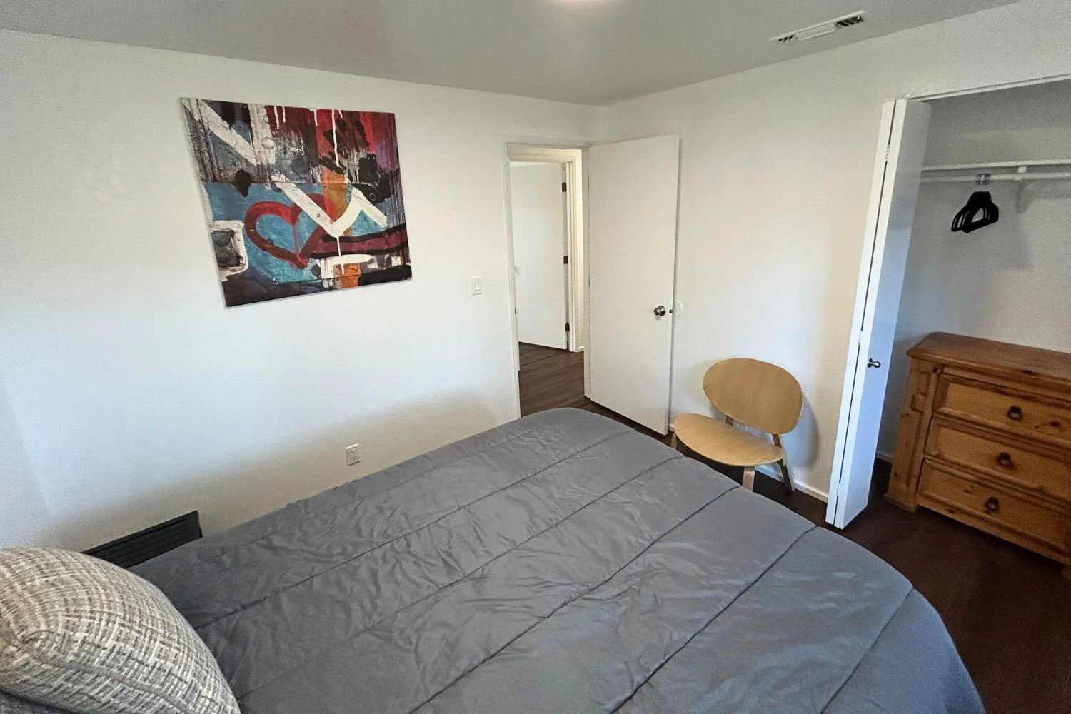 A bedroom with a bed, a chair, a dresser, an open closet, and a colorful abstract painting on the wall.