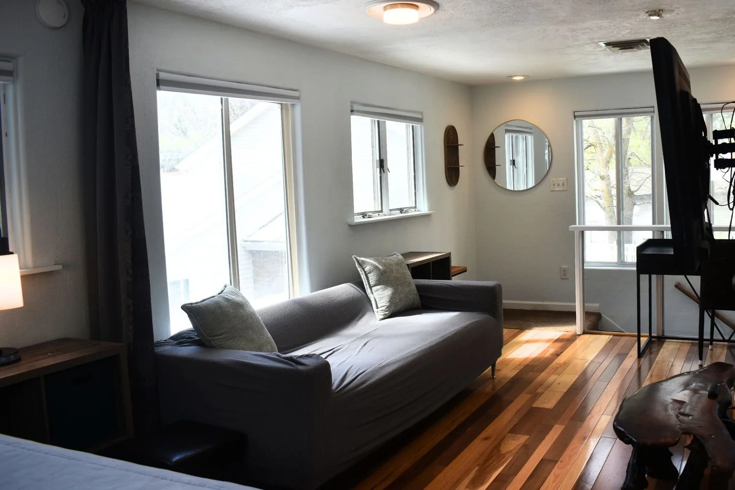 Living room with a gray sofa, wooden floors, multiple windows, a round mirror, and a small desk.