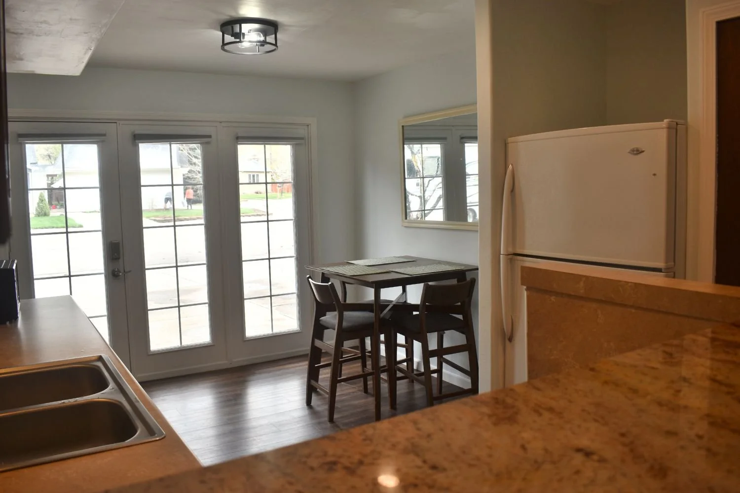 Kitchen area with granite countertop in the foreground, white refrigerator on the right, a small table with two chairs near large glass doors, and a mirror on the wall reflecting the outside view of a street and houses.