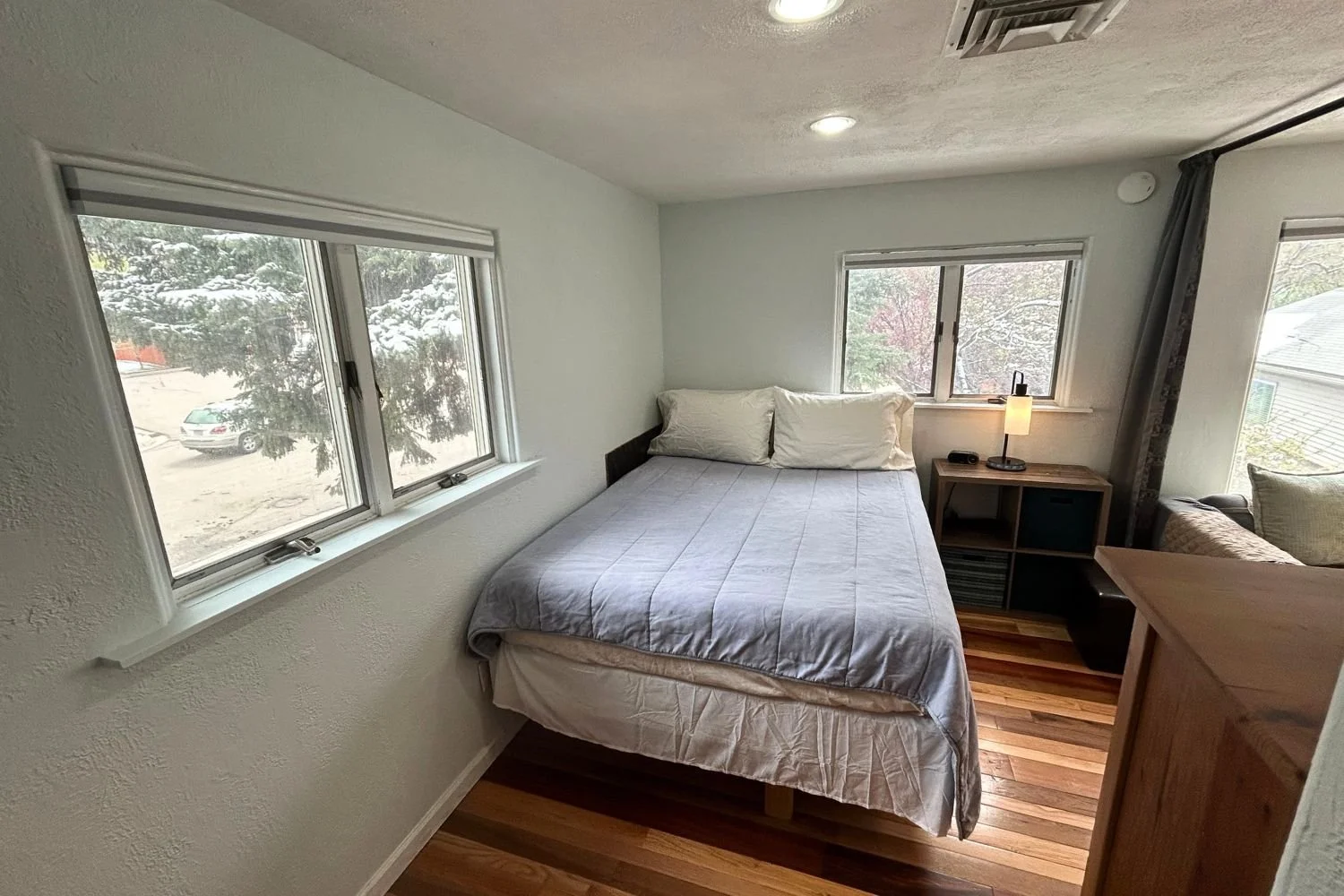Small bedroom with a bed, two pillows, a nightstand with a lamp, and windows showing snow outside.
