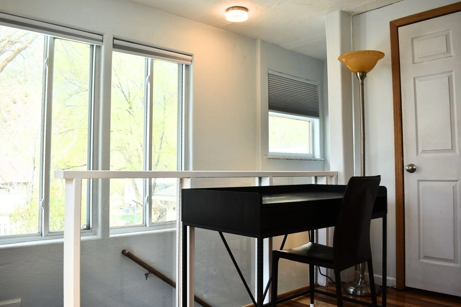 Interior of a room with large windows, a black desk with a chair, a white railing, a wooden floor, a tall floor lamp, a small window with blinds, and a white door.