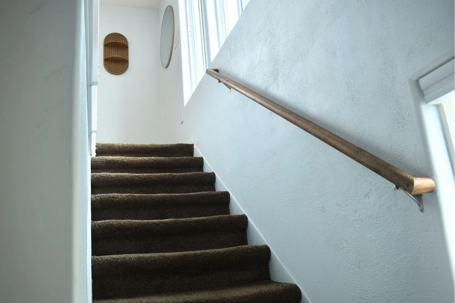 Indoor staircase with brown carpeted steps, a wooden handrail on the right, a small oval mirror on the wall, and a decorative wall shelf, with windows providing natural light.