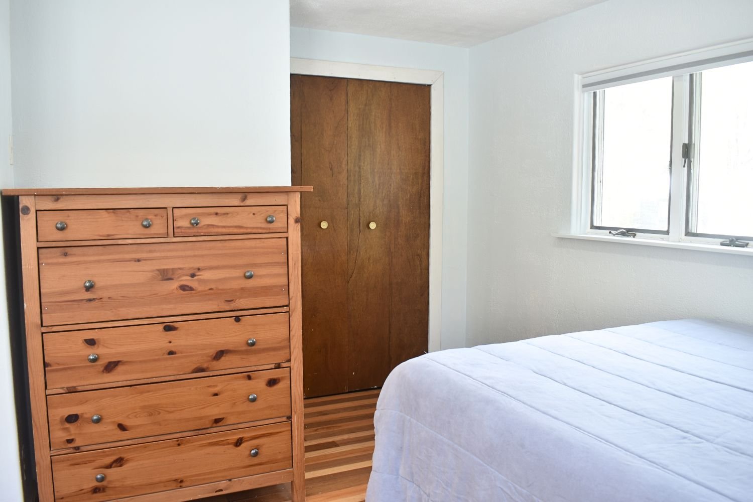 A bedroom with a wooden dresser, closet doors, a window with blinds, and part of a bed with a white quilt.