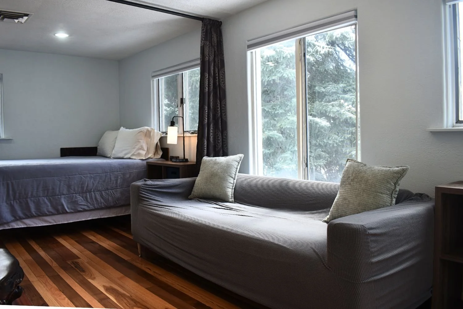 A living space with a bed on the left and a gray sofa with throw pillows on the right, near large windows with trees outside, hardwood floors, and a side table with a lamp.