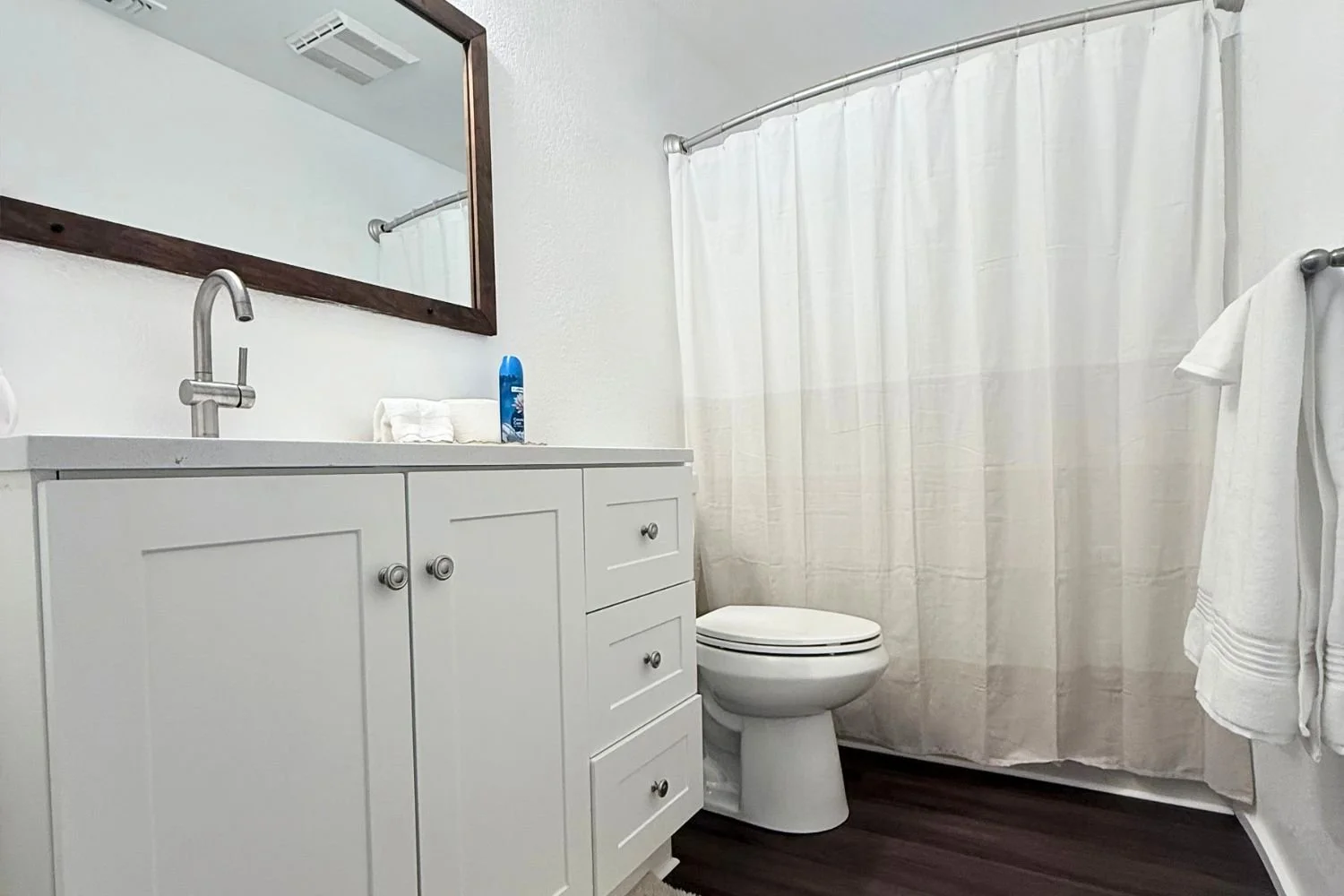 A small bathroom with white walls, a white vanity with silver knobs, a mirror with a wooden frame, a toilet, a white shower curtain, and a towel hanging on a silver rod. The bathroom has dark wooden flooring.