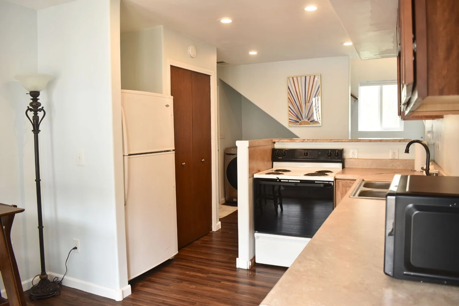 A kitchen with white walls, a refrigerator, an electric stove, a microwave, a sink, and wooden cabinets. There is a lamp on the left and a window on the right.