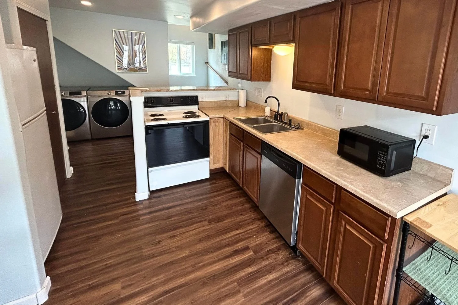 A kitchen with brown wooden cabinets, a beige countertop, a stainless steel dishwasher, a black microwave, a double sink, a white stove, and a laundry area with a washing machine and dryer in the background.