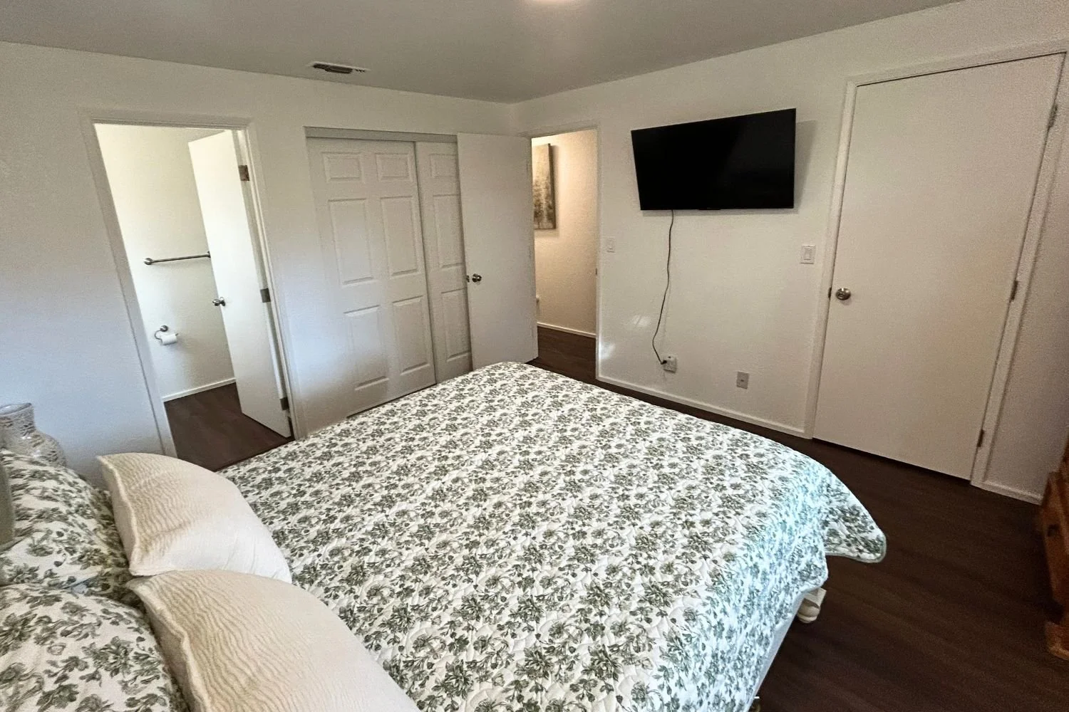 A bedroom with a bed covered in floral bedding, a wall-mounted TV, a closet with double doors, an open door leading to a hallway, and a bathroom with a toilet visible.