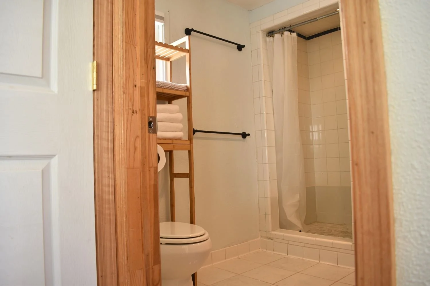 A bathroom with a shower, toilet, and wooden shelving with towels, seen through a partially open door.