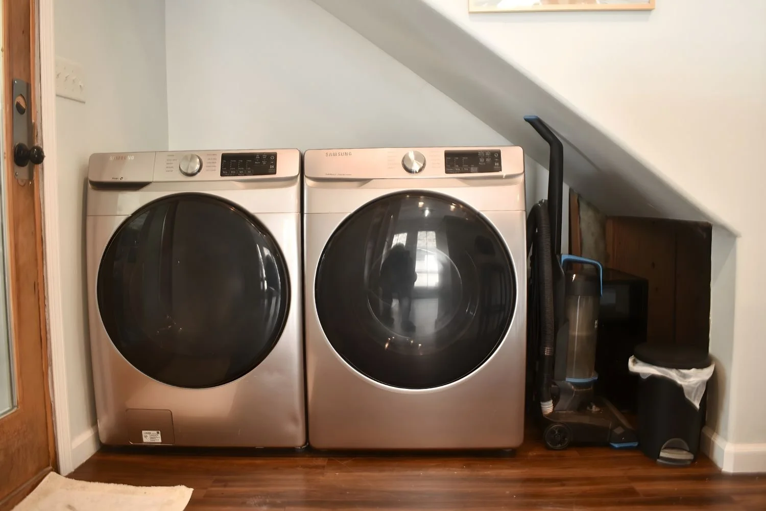 A laundry room with a side-by-side washer and dryer, both in silver color, situated under a sloped ceiling. To the right, there's a black and blue vacuum cleaner, a small black shelving unit, and a trash can covered with a plastic bag. The floor is w