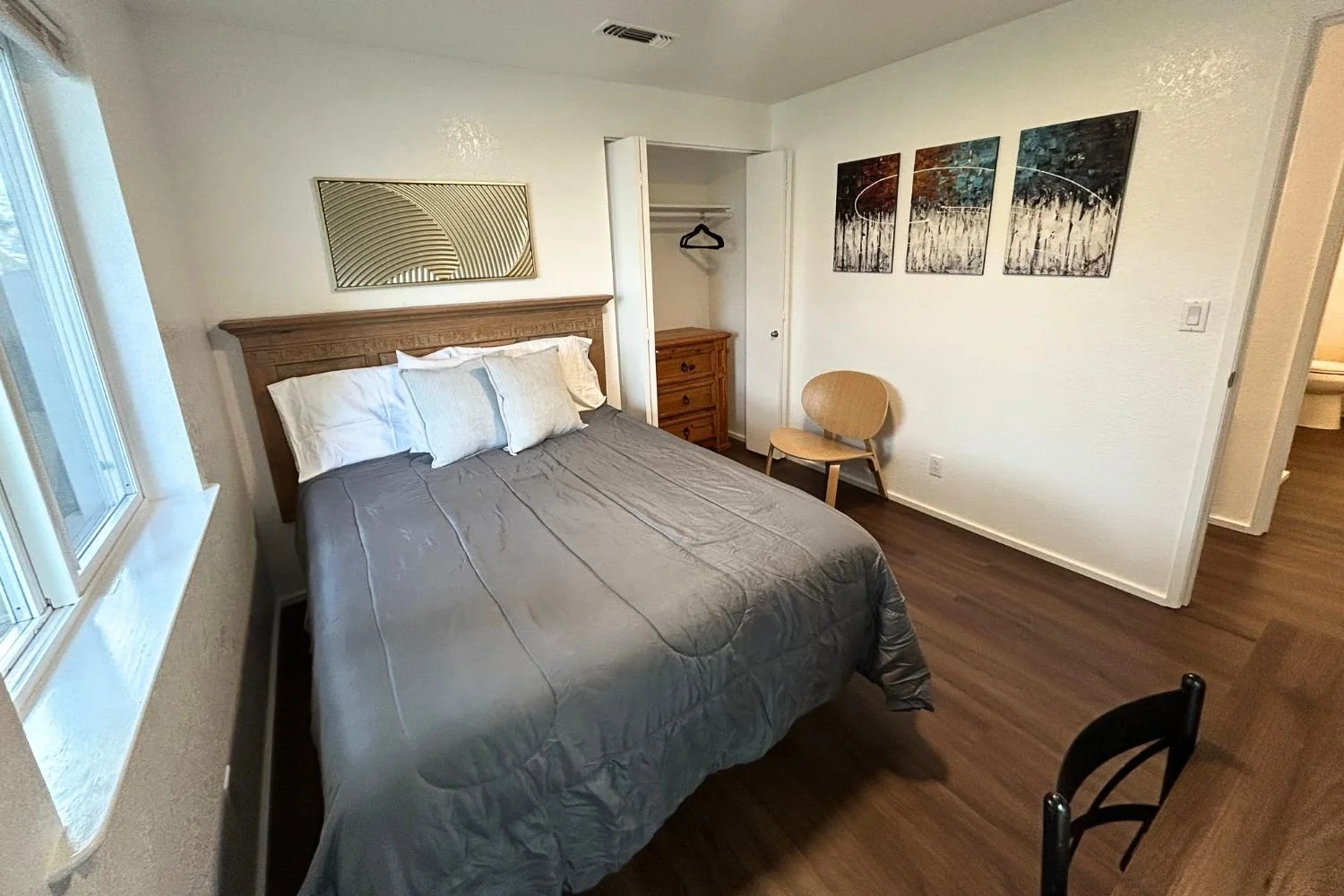 A bedroom with a bed, pillows, a window, a wooden chair, a small wooden nightstand, a closet with an open door, and three abstract paintings on the wall.