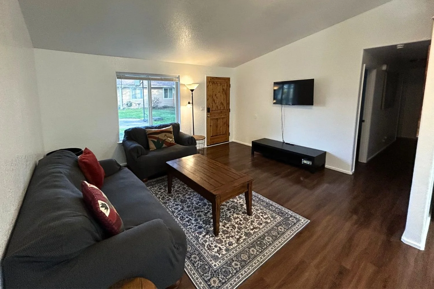 Living room with two dark-colored sofas, a wooden coffee table on an ornate rug, a wall-mounted TV, a window with a view of a yard, and a wooden door.