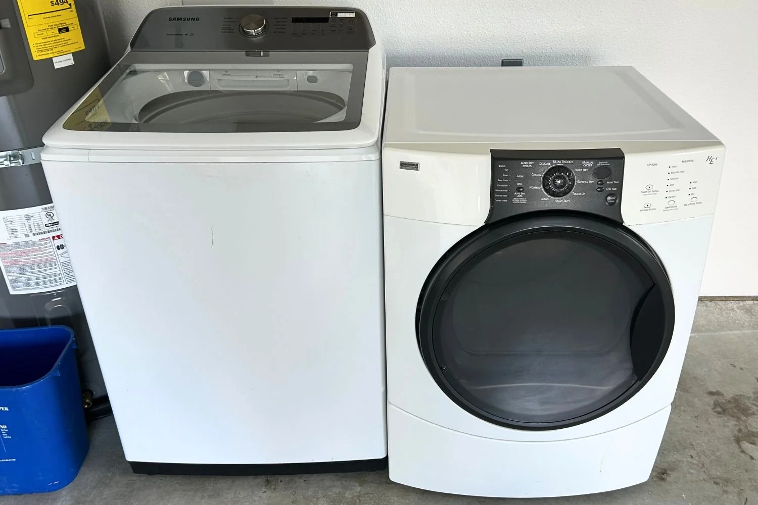 A side-by-side of a top-loading washing machine on the left and a front-loading dryer on the right, both in white, in a laundry room setting.