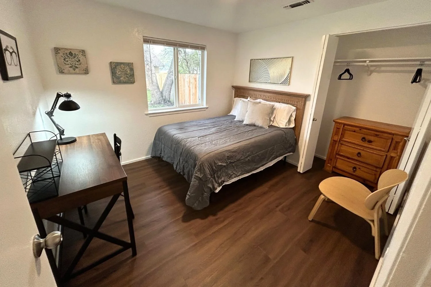A small bedroom with a bed, desk, chair, dresser, and closet, featuring hardwood flooring and a window overlooking a backyard.