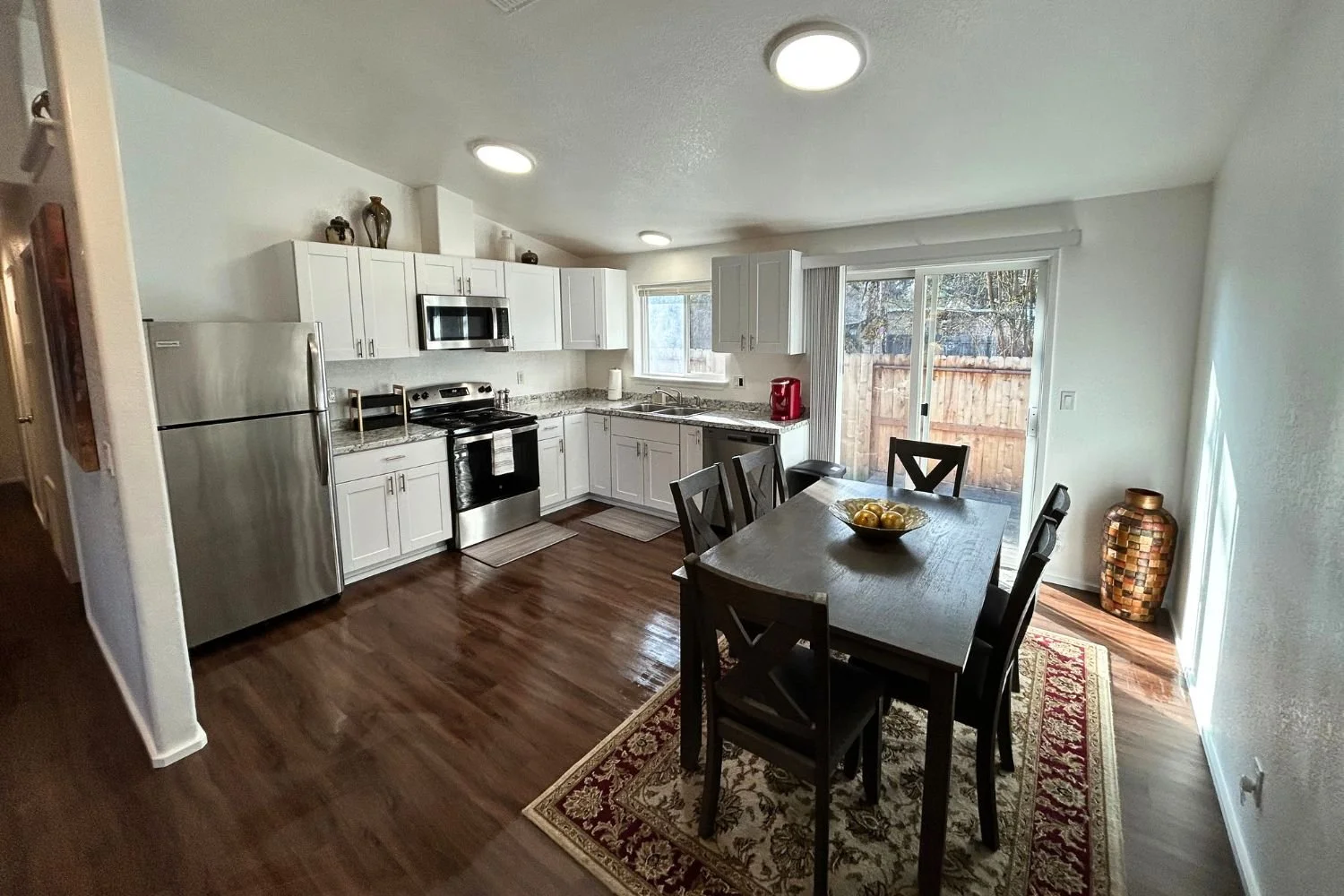 A kitchen with white cabinets, stainless steel refrigerator, stove, microwave, and granite countertops. There is a dining table with six chairs and a sliding glass door to the backyard. The room has wooden flooring and a decorative vase in the corner