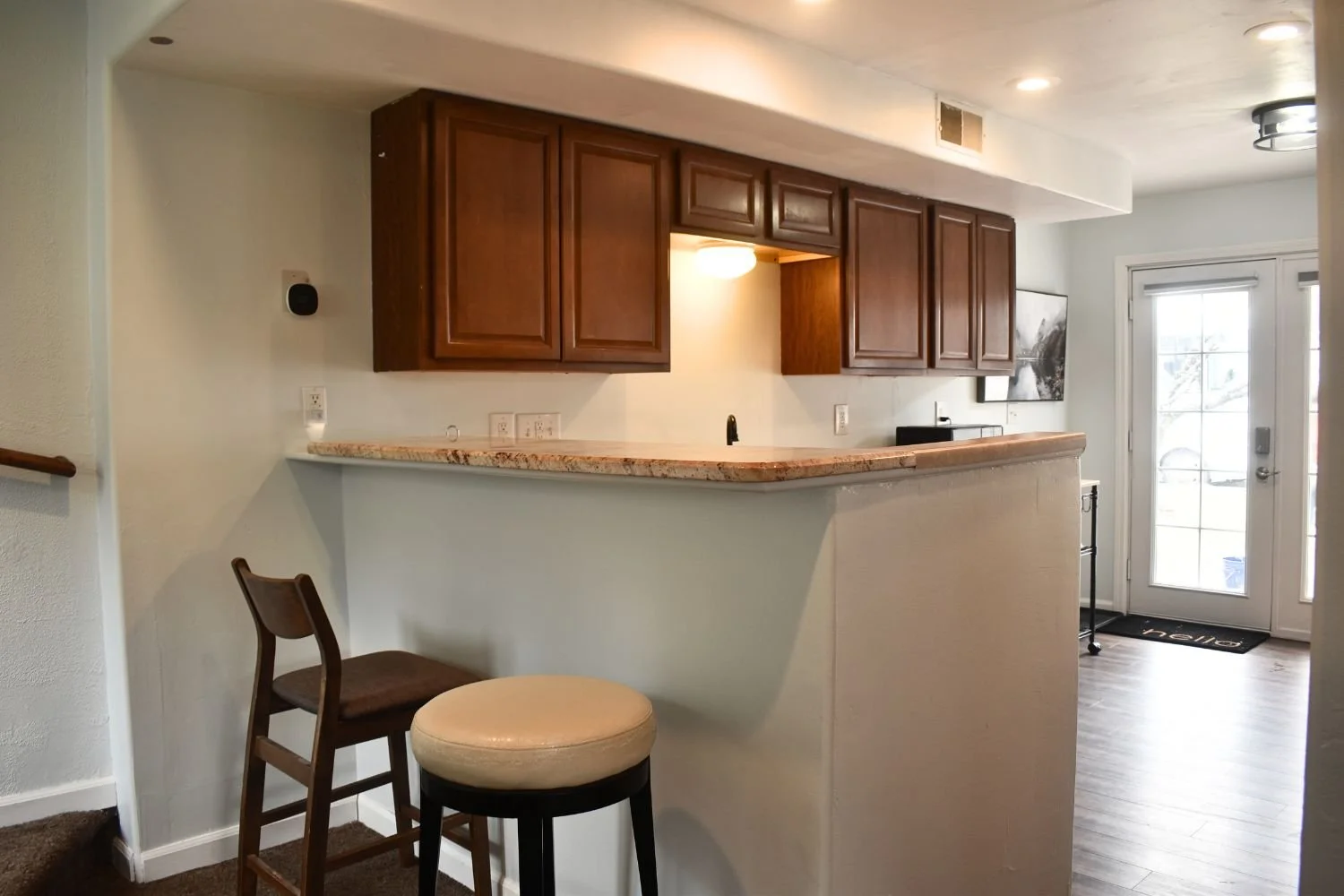 Interior view of a kitchen with brown cabinets, granite countertop, and a partial wall with two bar stools in the foreground. There is a door with glass panes leading outside and a painting on the wall.