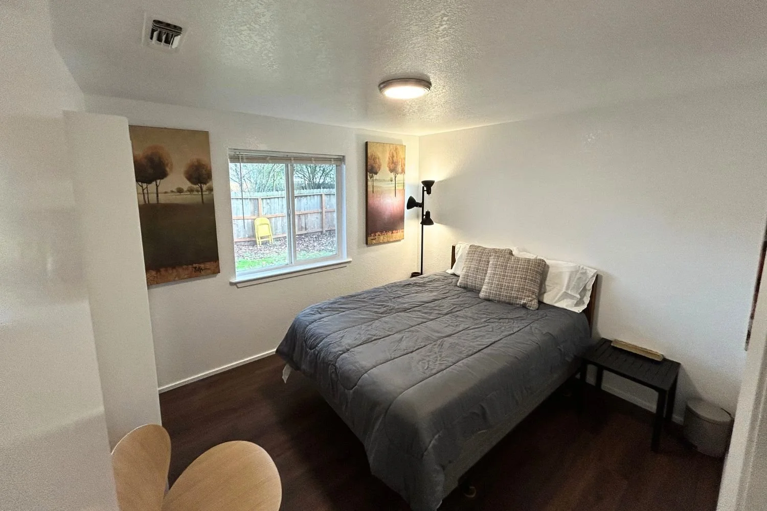 A bedroom with a bed, two pillows, a small side table with a book, wall art, a window showing an outdoor yard, and a floor lamp.