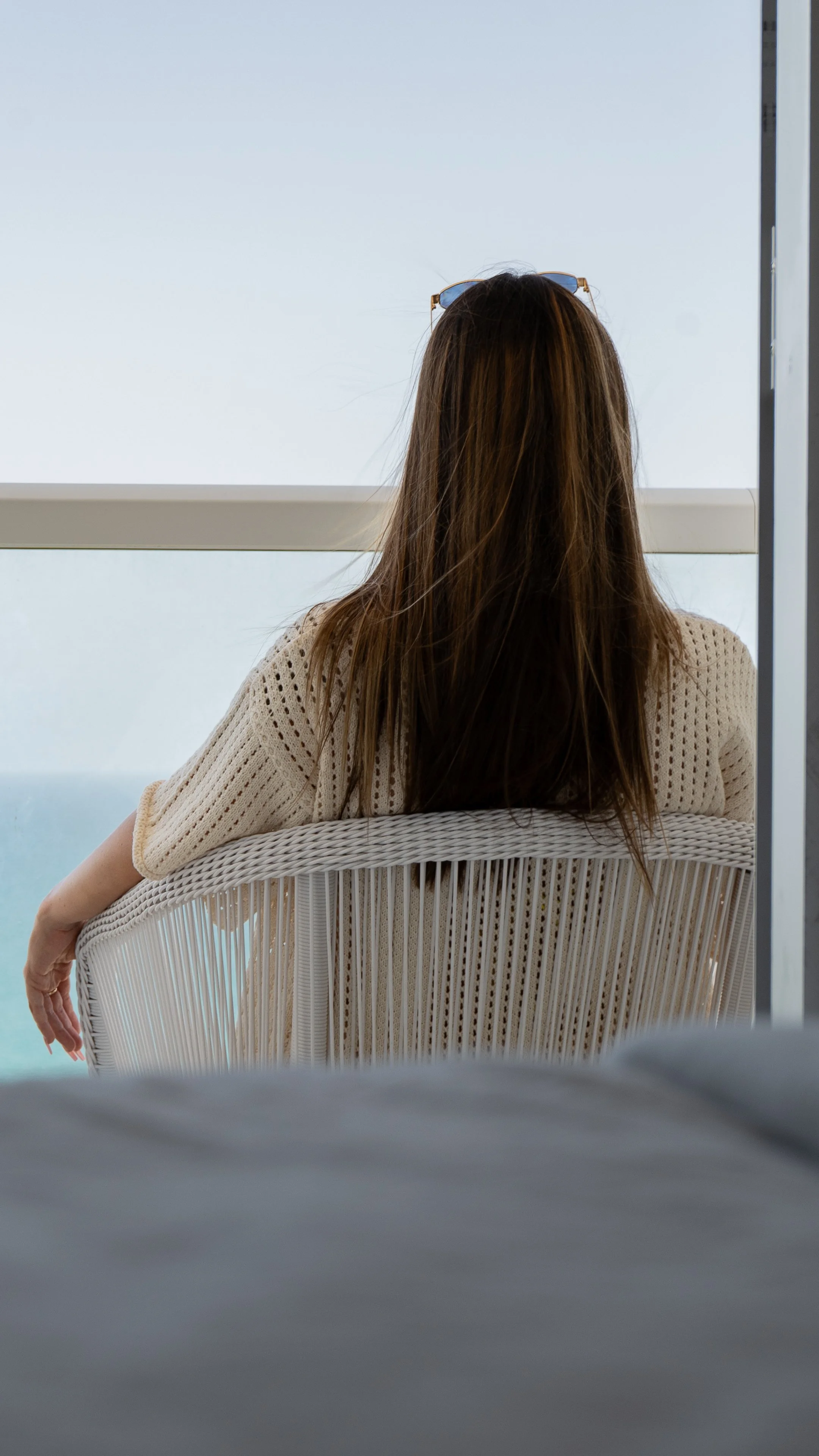 A woman with long brown hair sitting on a white wicker chair, looking out through a large window over a body of water, with sunglasses resting on her head and wearing a cream-colored knit top.