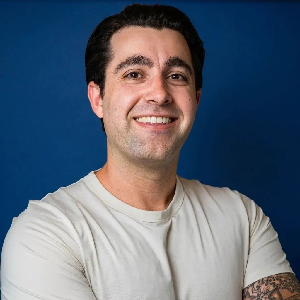 A smiling man with dark hair and a tattoo on his left arm, wearing a light-colored t-shirt, standing in front of a blue background.