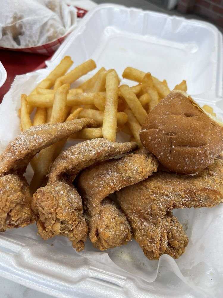 Fried chicken wings with a dusting of powdered seasoning, French fries, and a small bread roll in a takeout container.