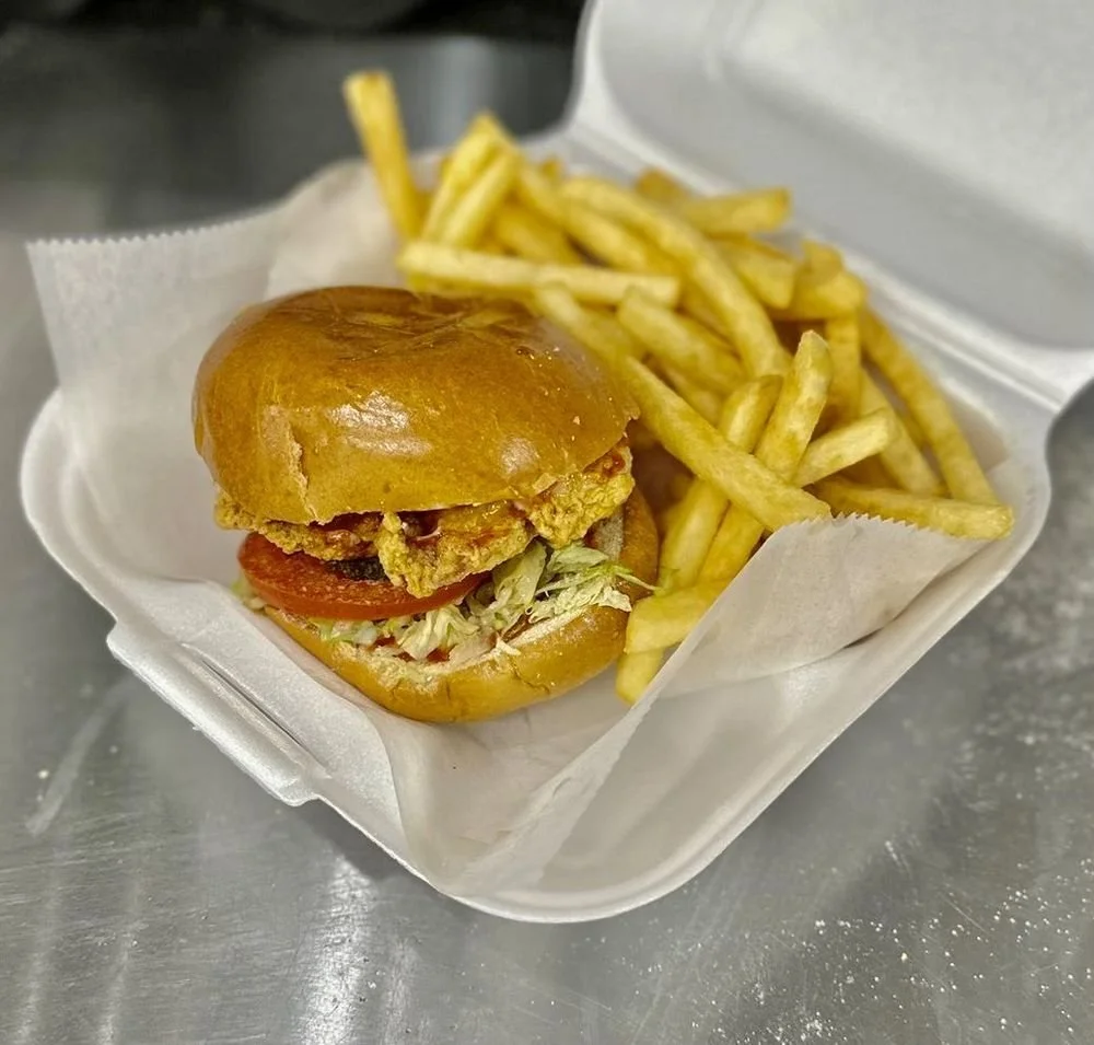 Fried chicken sandwich with lettuce, tomato, and sauce in a bun, served with French fries in a white takeout container.