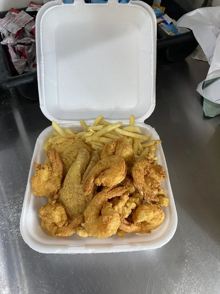 A foam takeout container with fried chicken pieces, French fries, and a fried fish fillet inside, placed on a metallic surface.