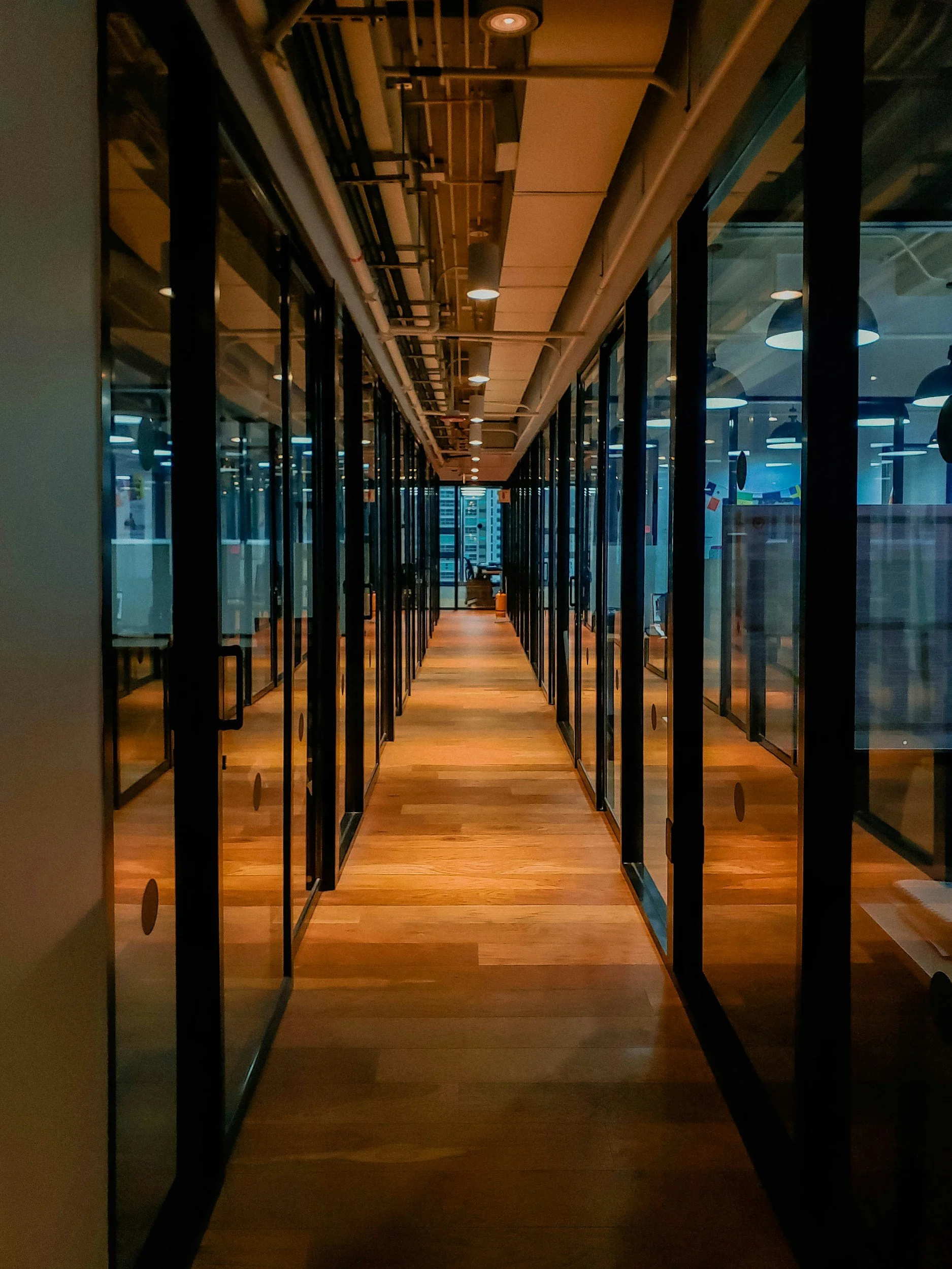 A modern office corridor with glass walls and wooden flooring, illuminated by ceiling lights, extending into a distant room.