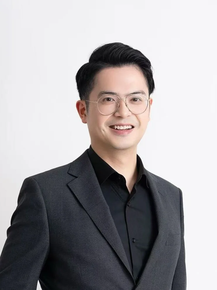 A young man with black hair, glasses, wearing a dark gray blazer and black shirt, smiling against a white background.