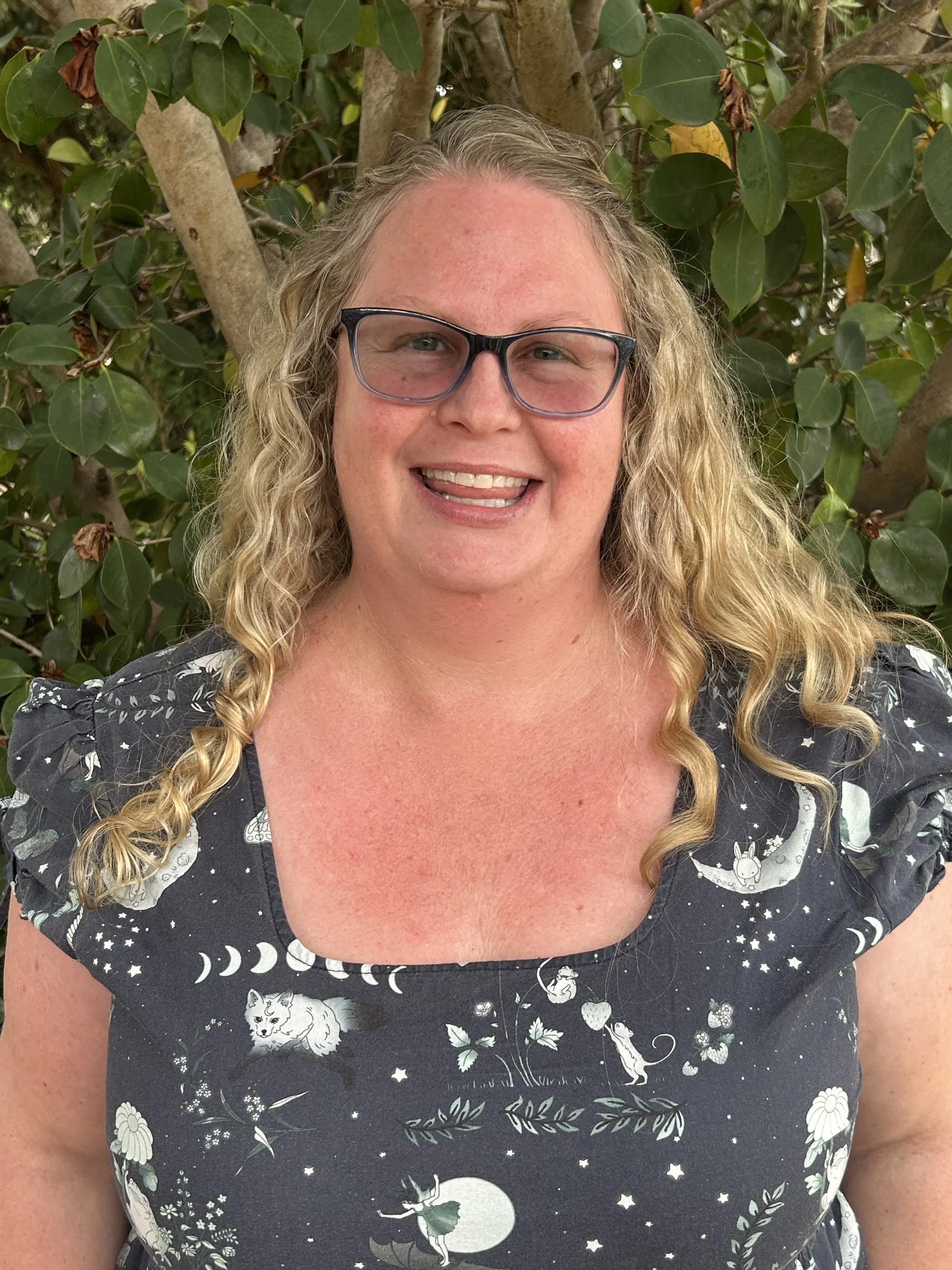 A woman with curly blonde hair, wearing glasses, smiling, standing outdoors against a background of green leaves and tree branches.