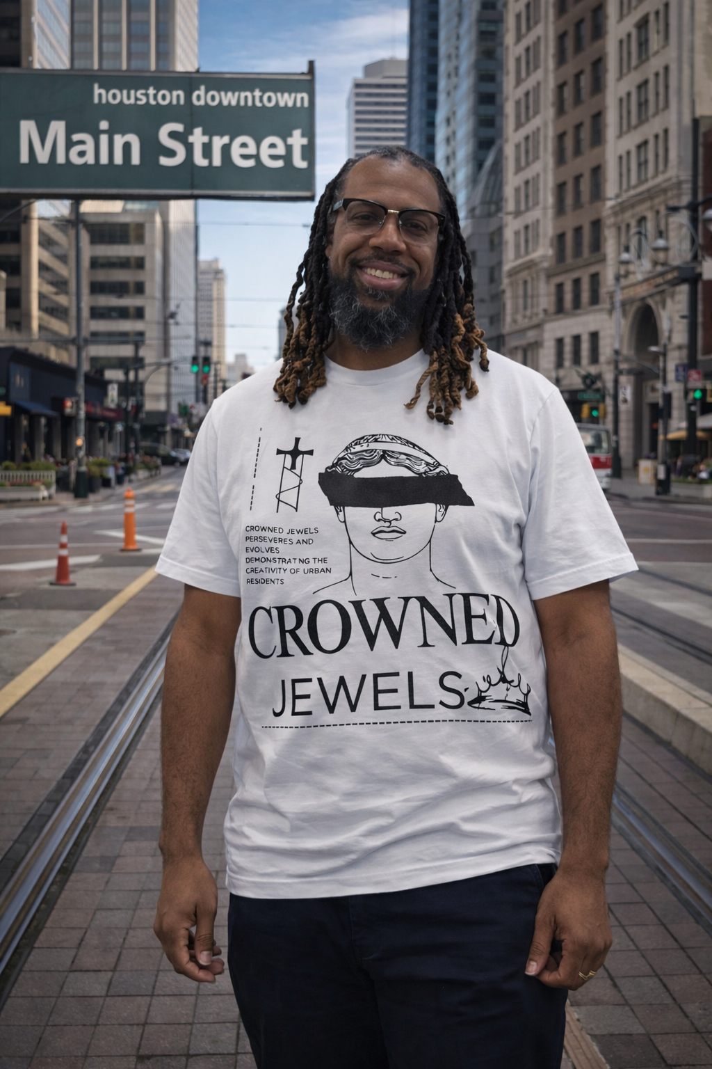 Smiling man with dreadlocks and glasses standing on Main Street in downtown Houston, wearing a white T-shirt with a graphic and text related to Crowned Jewels, in an urban setting with buildings and traffic.