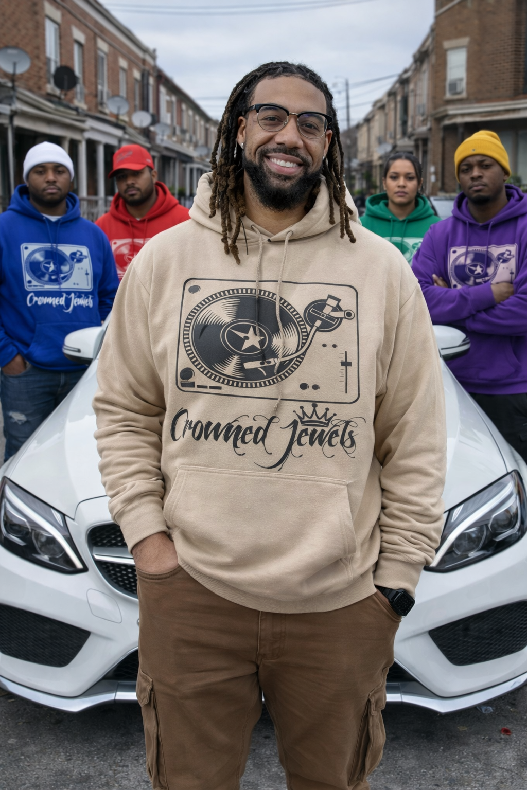 A man with glasses and dreadlocks smiling in front of a white car, wearing a beige hoodie with a DJ graphic and 'Crowned Jewels' text; four people in colorful hoodies stand behind him on a city street.